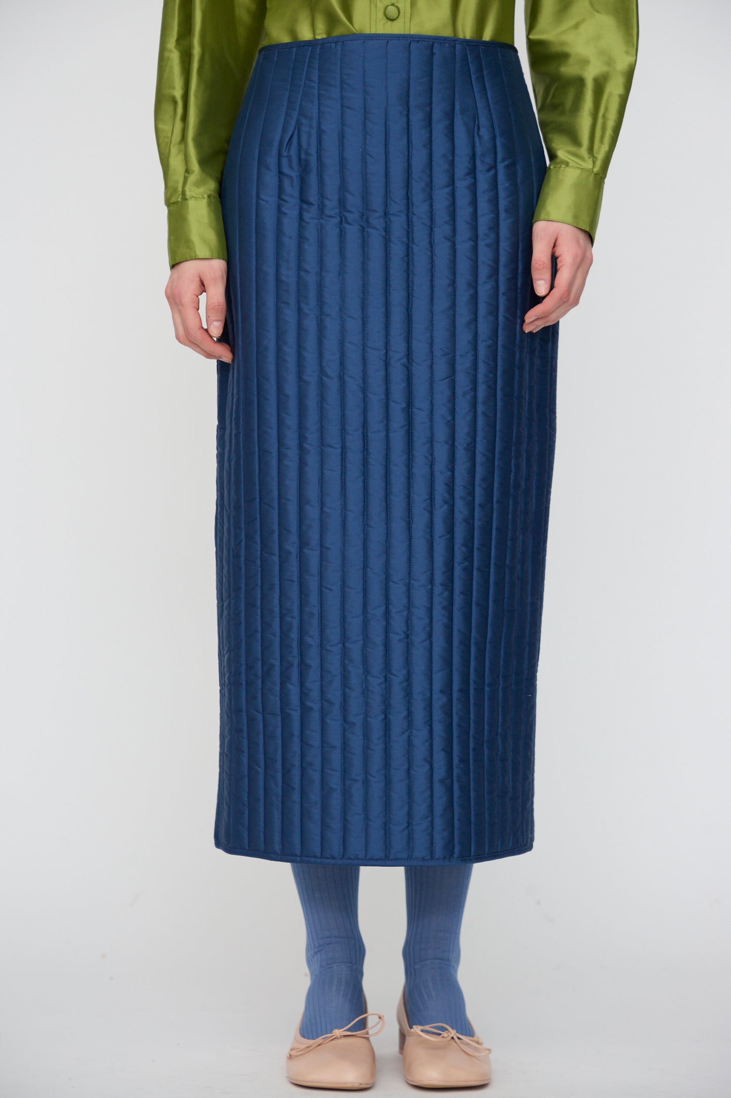 A person wearing a green satin long-sleeve shirt, Cawley's Quilted Silk Sadie Skirt in Insignia, blue socks, and beige ballet flats stands against a plain white background.