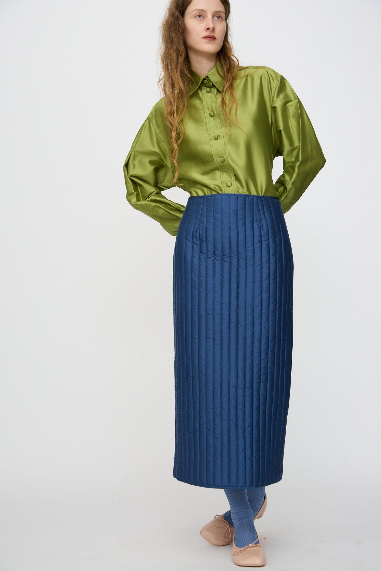 A person wears a shiny green button-up shirt, blue tights, light pink shoes, and the Cawley Quilted Silk Sadie Skirt in Insignia while standing against a plain white background.