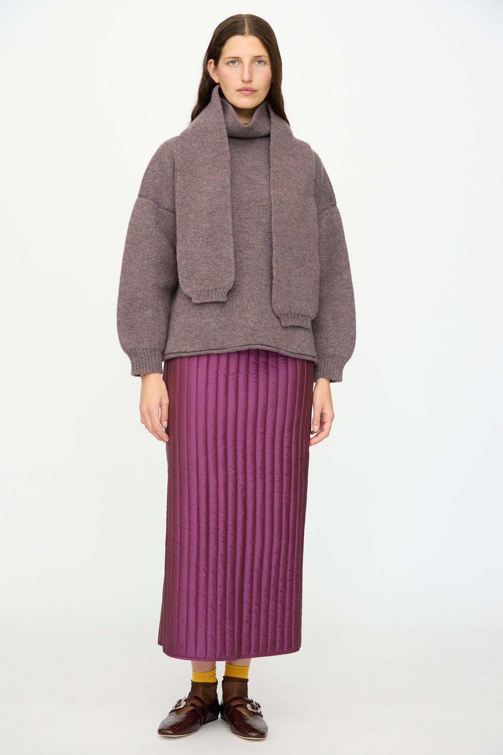 A woman faces forward wearing a mauve sweater with a matching scarf, the Cawley Quilted Silk Sadie Skirt in Plum, yellow socks, and brown shoes against a plain white background.