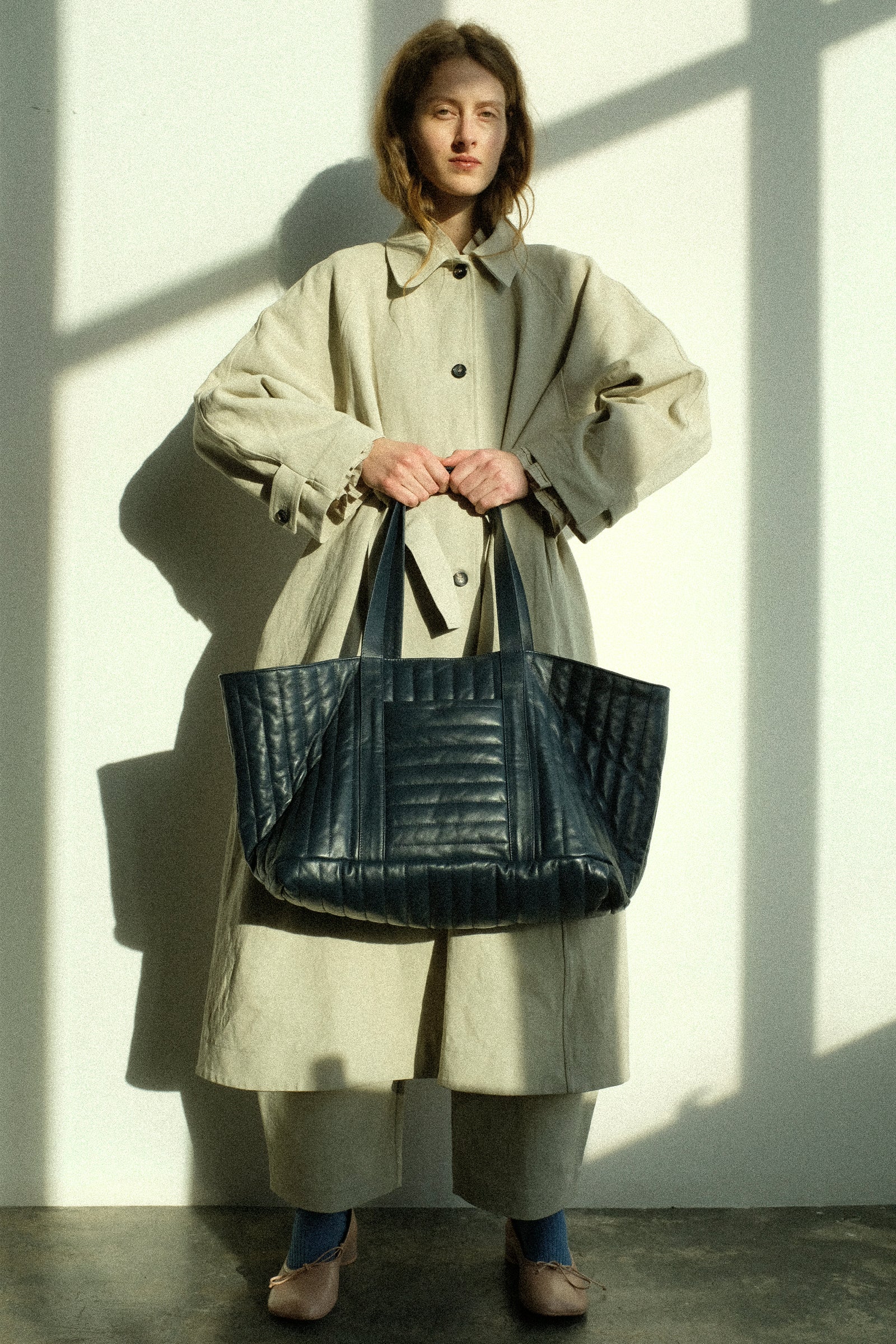 Person wearing the Japanese Drill Wickham Mac in Natural from Cawley holding the Quilted Nappa Large Tote Bag with Coin Purse in Navy against a light background