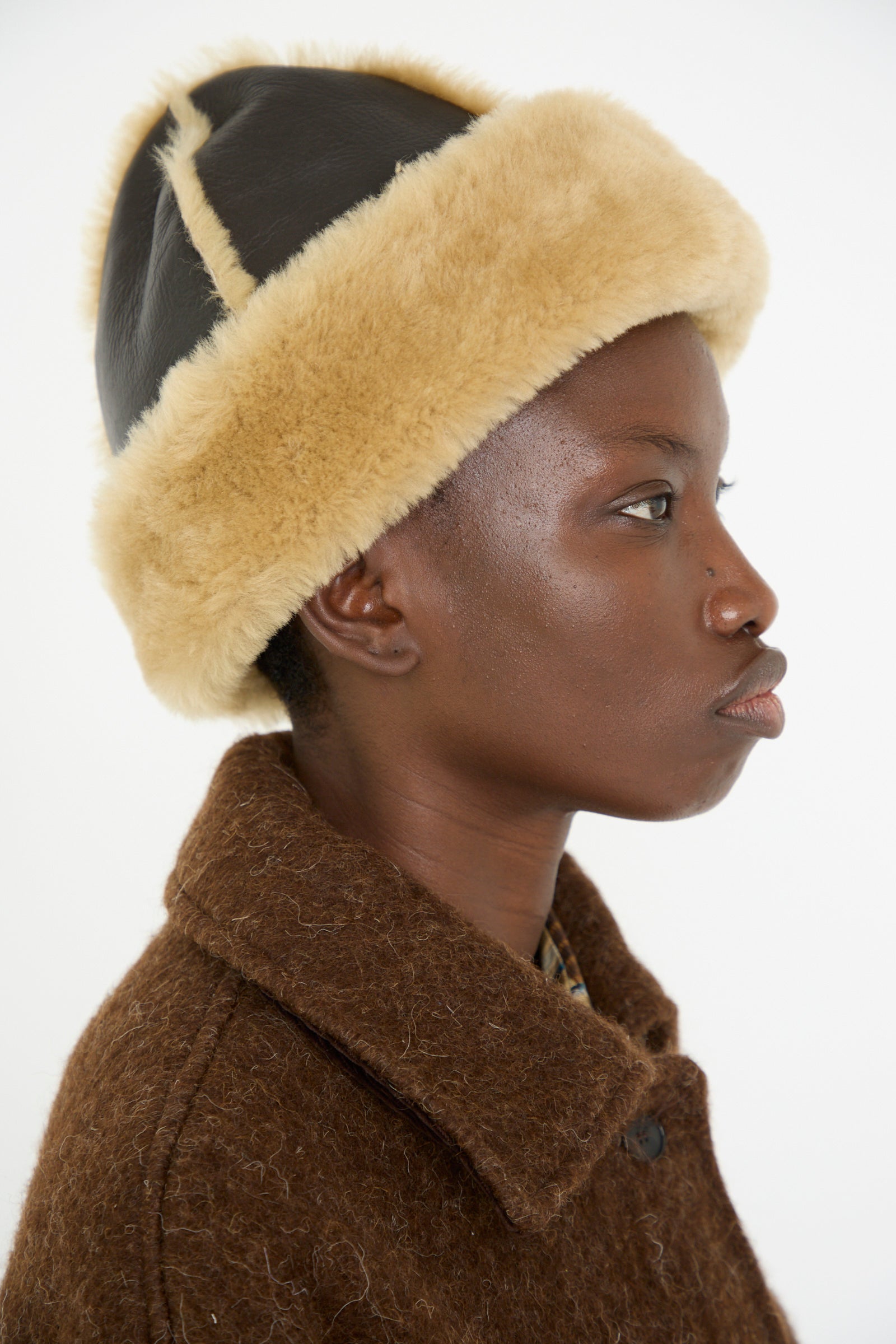 A person wearing a brown coat and a Chocolate and Tan Sheepskin Leather Seam Hat by Cawley, made in London, is shown in profile against a plain white background.