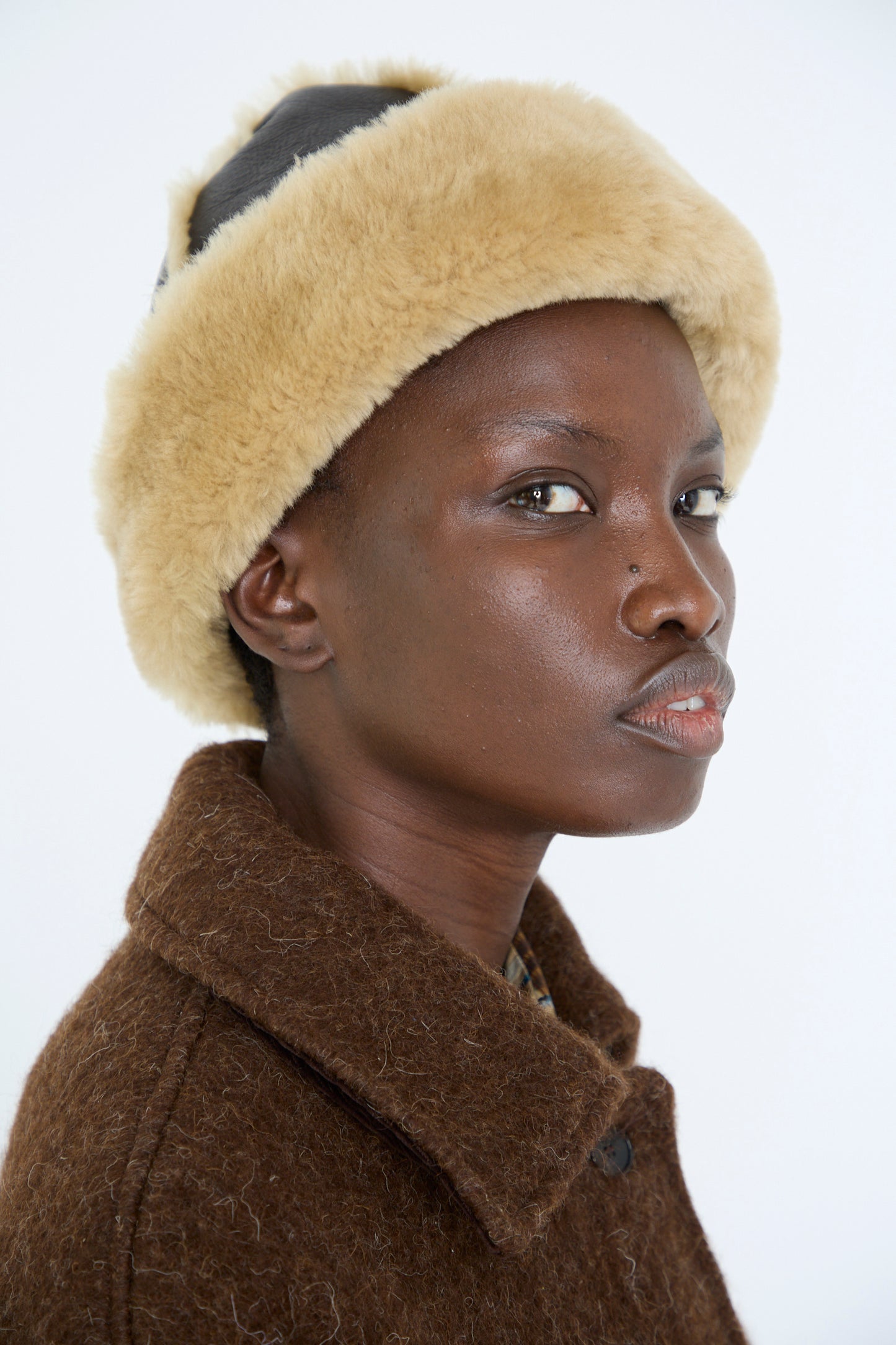 Dressed in a chocolate and tan Sheepskin Leather Seam Hat by Cawley, a person wearing a brown wool coat glances to the side against a plain background.