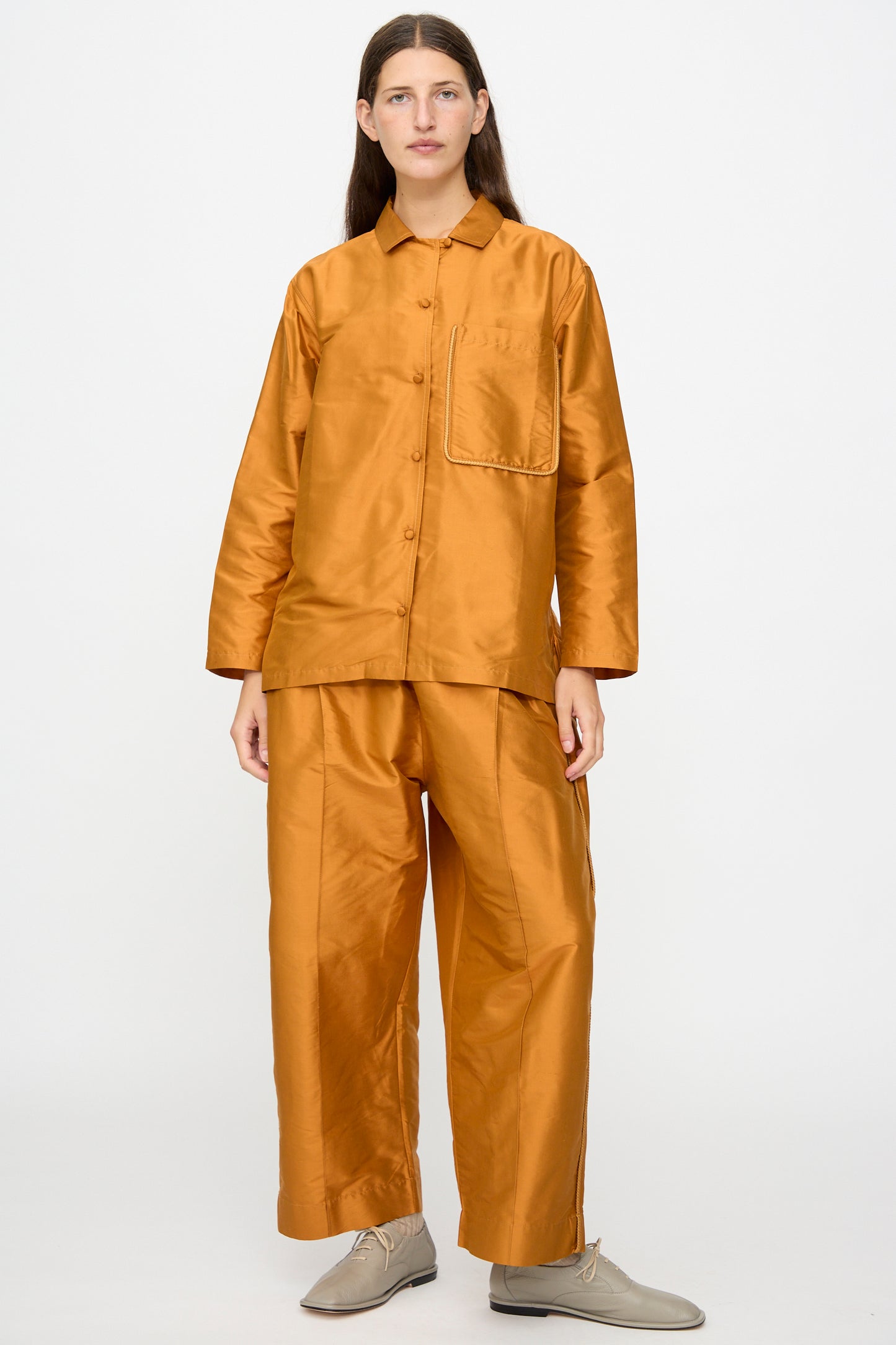 A person stands against a plain background, wearing the Cawley Silk Dupion August Shirt in Sienna with matching wide-leg pants and beige lace-up shoes.