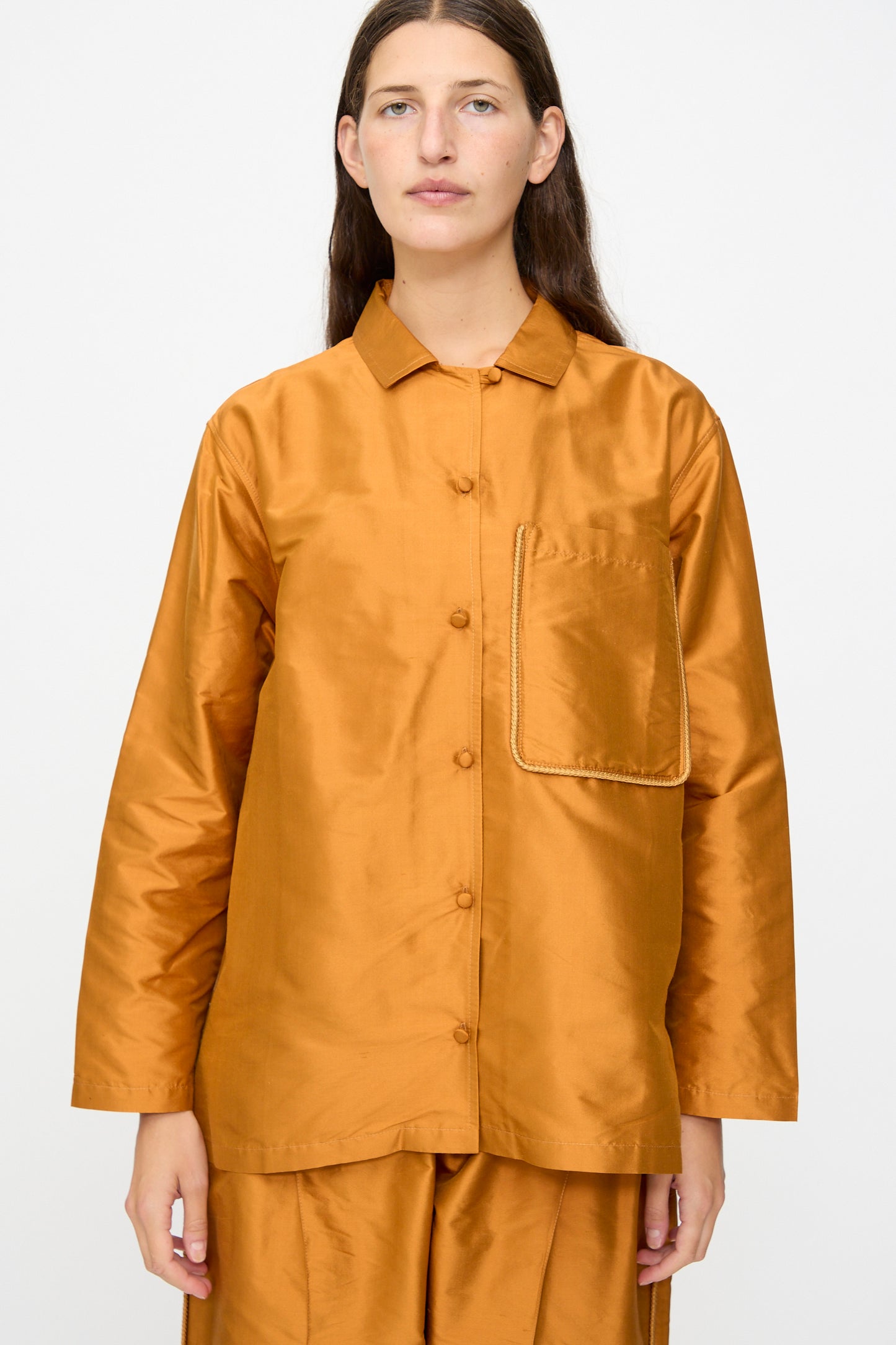 A person wearing the Cawley Silk Dupion August Shirt in Sienna—a shiny, loose-fitting silk shirt with long sleeves and a large front pocket—paired with matching pants, stands against a plain background.