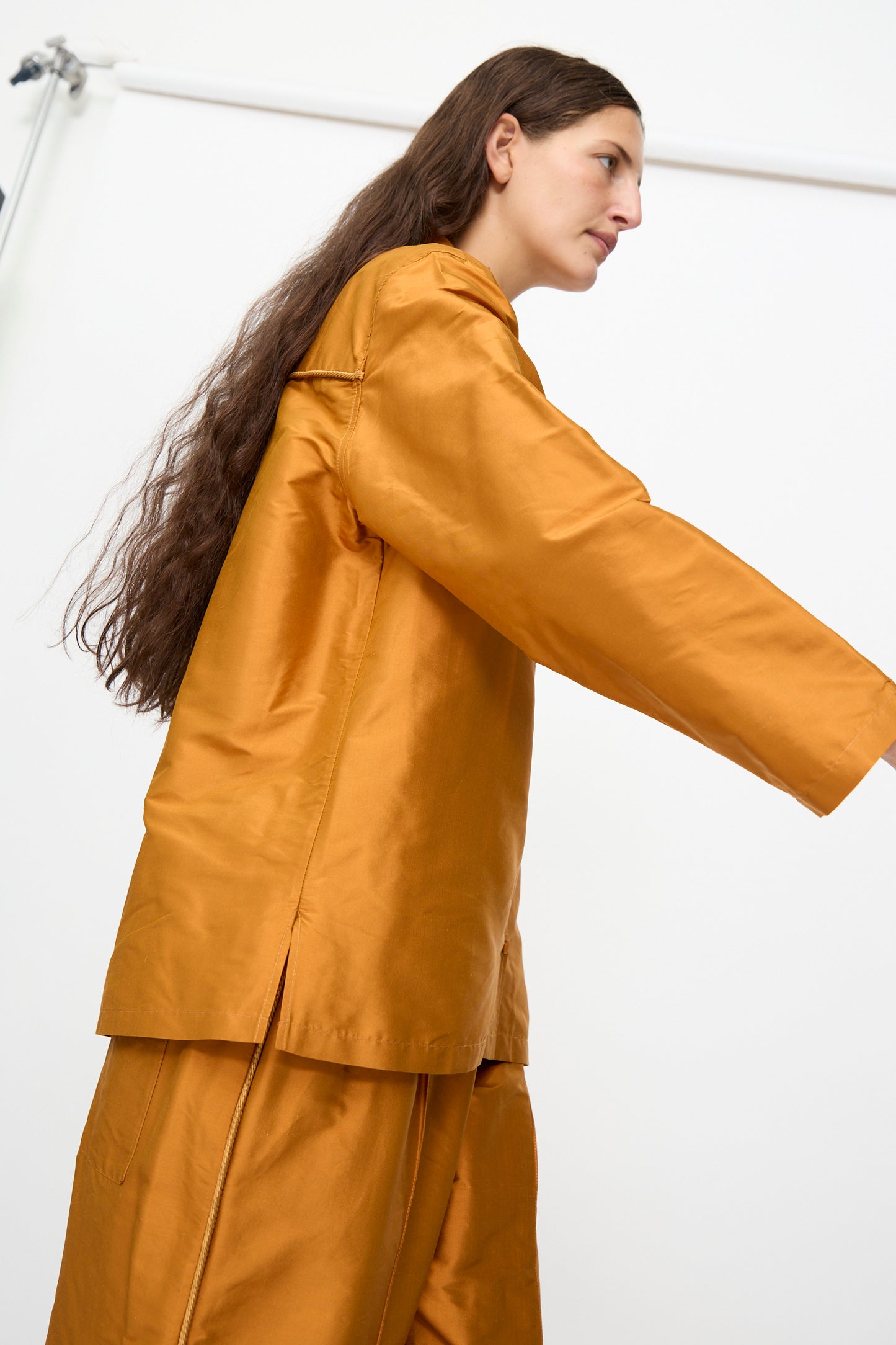 Someone with long brown hair wears the Cawley Silk Dupion August Shirt in Sienna, a loose-fitting, golden-brown silk shirt made in the UK, standing sideways against a plain white background.