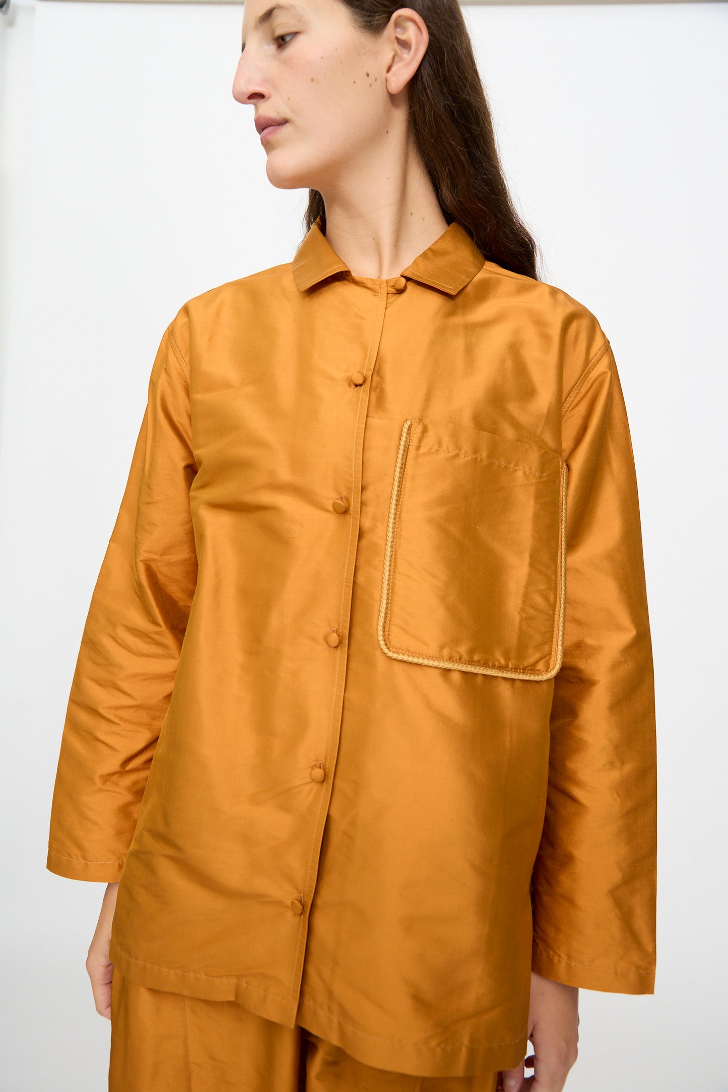 A person wearing the Cawley Silk Dupion August Shirt in Sienna—a loose-fitting, long-sleeved, copper-brown silk shirt with a large front pocket, made in the UK—stands against a plain white background.