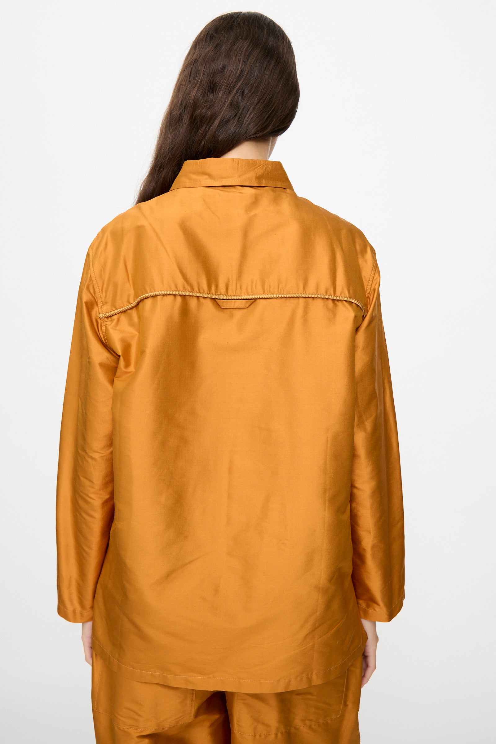 A person with long brown hair is photographed from the back against a plain white background, wearing the Cawley Silk Dupion August Shirt in Sienna—a loose-fitting, long-sleeved shirt crafted in the UK—and matching pants.