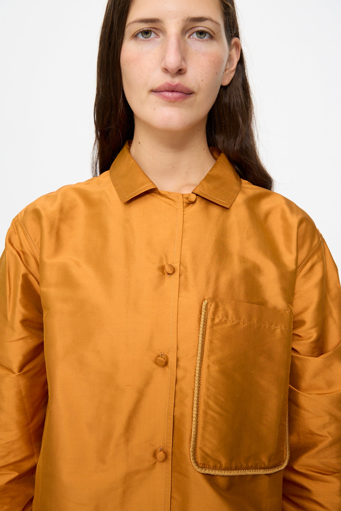 A person with long brown hair wears the Cawley Silk Dupion August Shirt in Sienna, featuring a large pocket on the left chest, standing against a plain white background.