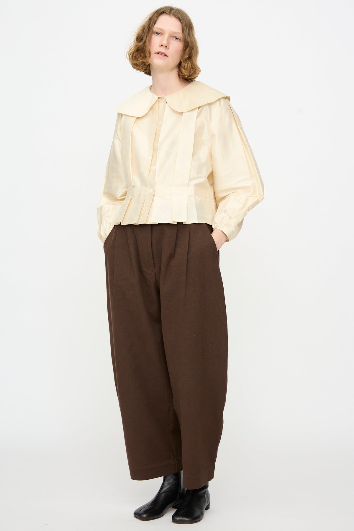 A person stands against a plain background wearing the Cawley Silk Dupion Box Pleat Blouse in Ivory, featuring a wide collar and long sleeves, styled with loose brown trousers and black ankle boots. Their hands are in their pockets.