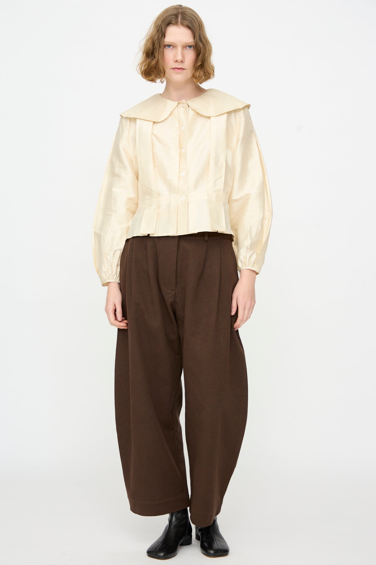 A person stands against a plain background wearing the Cawley Silk Dupion Box Pleat Blouse in Ivory with long sleeves and a wide collar, styled with brown wide-leg pants and black ankle boots. This elegant look is made in the UK.