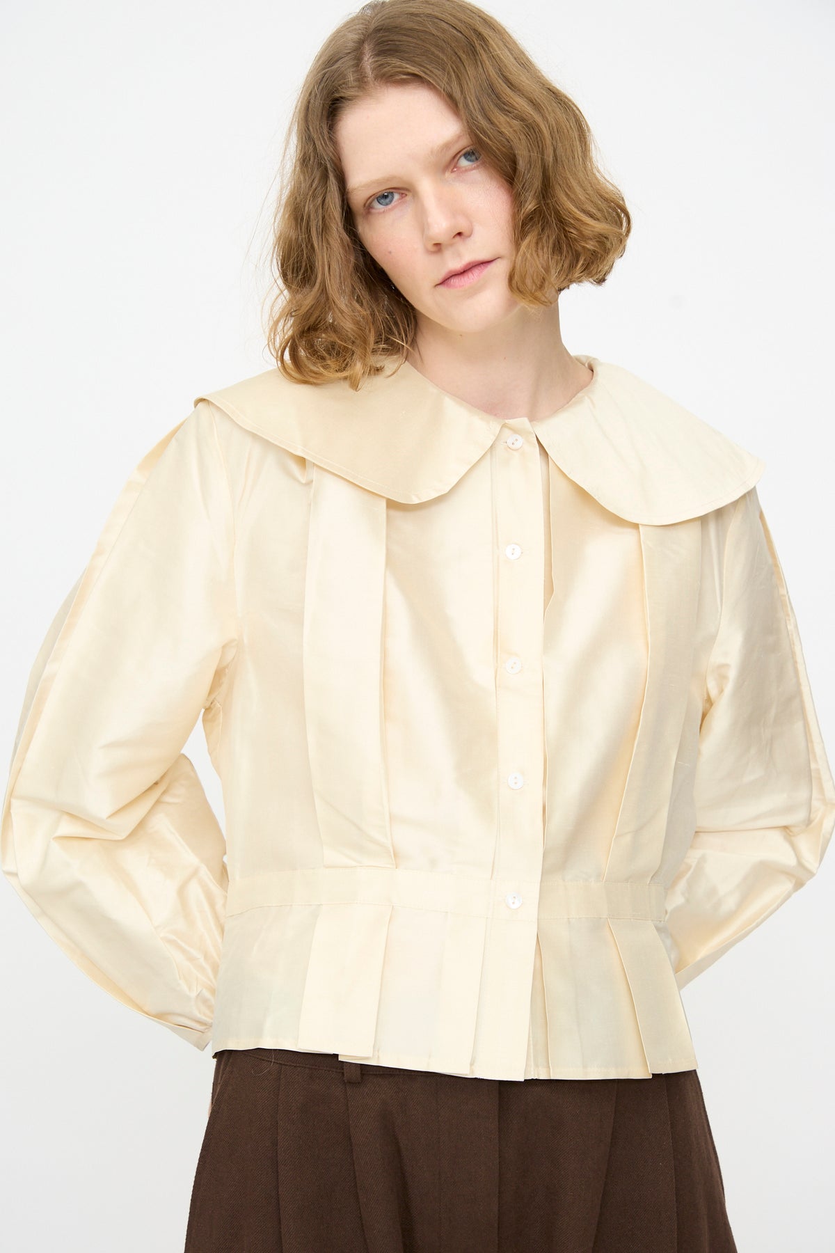 A woman wears the Cawley Silk Dupion Box Pleat Blouse in Ivory—an elegant, cream-colored, long sleeve blouse featuring a wide collar and pleated details—paired with brown pants and standing against a plain background. This sophisticated shirt is made in the UK.