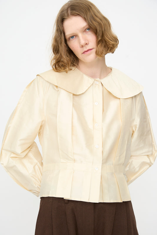 A woman wears the Cawley Silk Dupion Box Pleat Blouse in Ivory—an elegant, cream-colored, long sleeve blouse featuring a wide collar and pleated details—paired with brown pants and standing against a plain background. This sophisticated shirt is made in the UK.