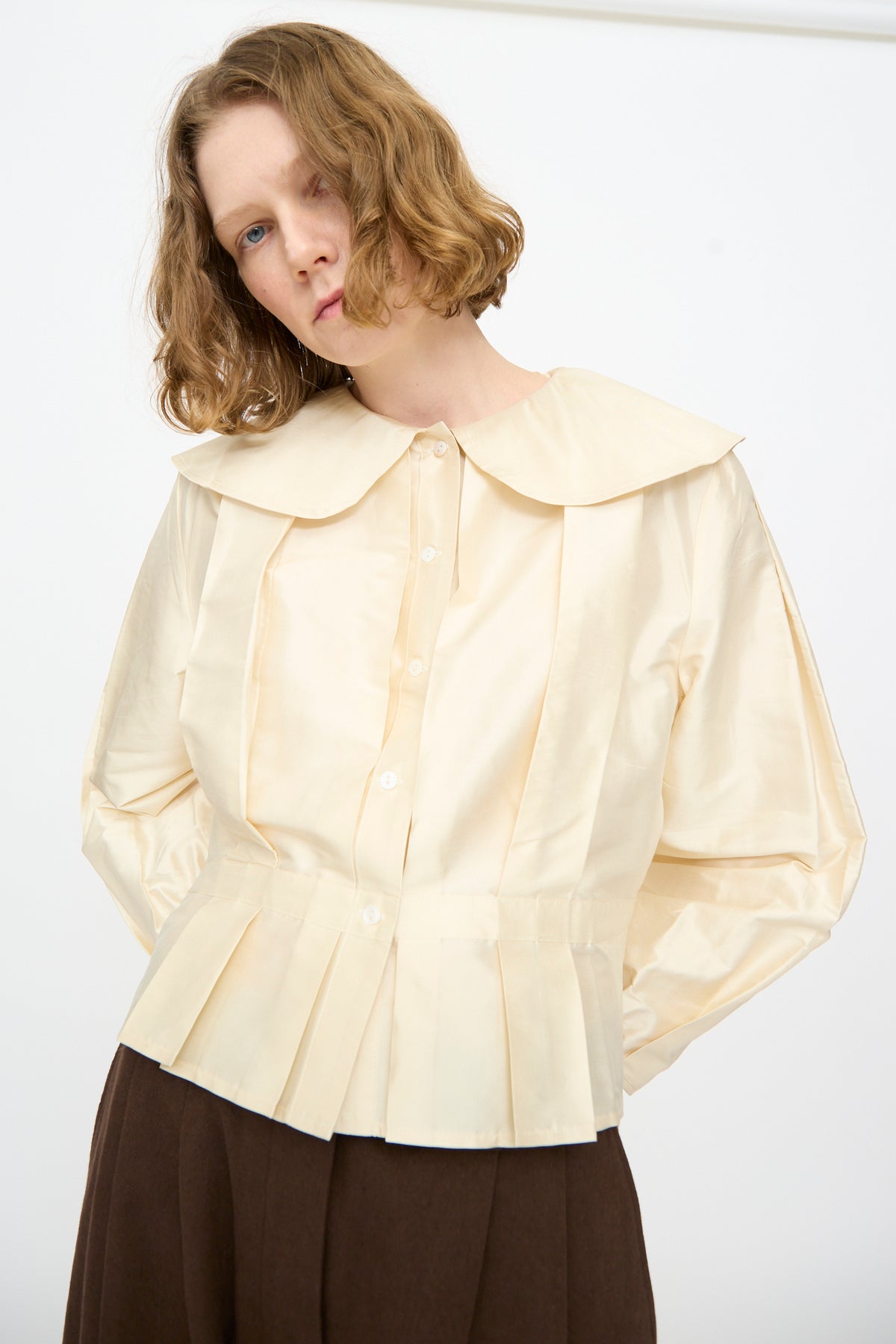 A person with short, wavy hair wears the Cawley Silk Dupion Box Pleat Blouse in Ivory—a long sleeve blouse featuring a large collar and pleated details—paired with brown pants, standing against a plain white background. This elegant piece is made in the UK.