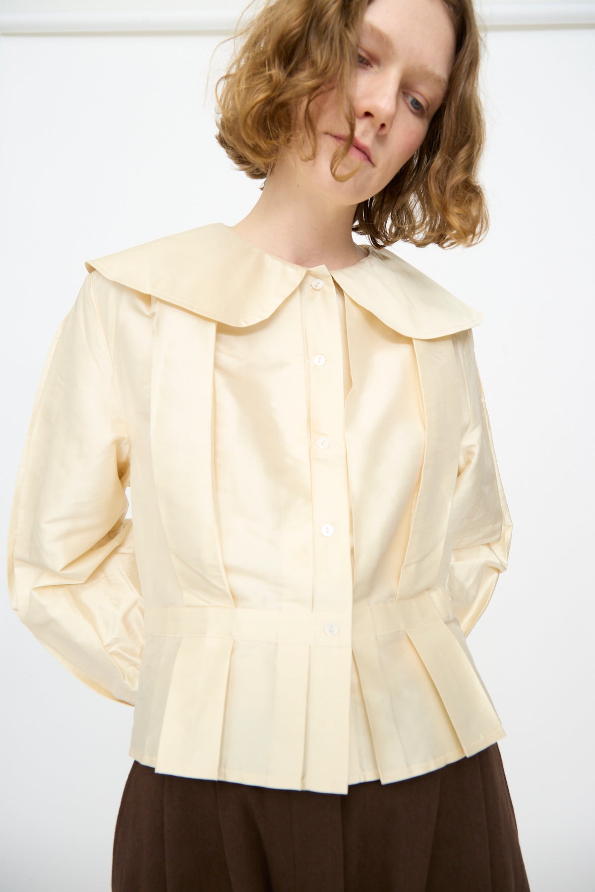 A person wearing the Cawley Silk Dupion Box Pleat Blouse in Ivory, featuring long sleeves, a wide collar, and pleated details, paired with brown pants and standing against a plain white background.
