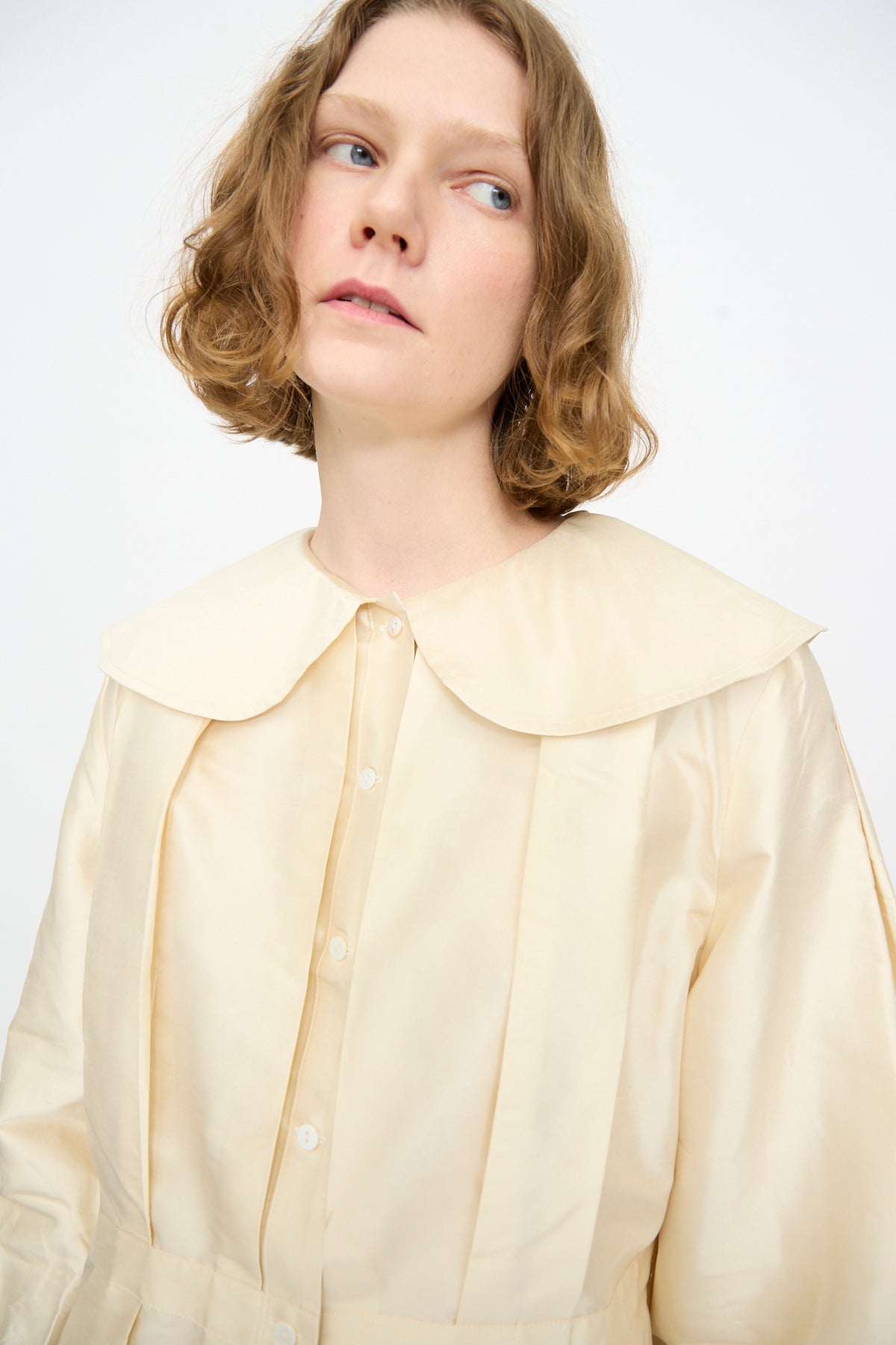 A person with short, wavy light brown hair, dressed in the Cawley Silk Dupion Box Pleat Blouse in Ivory, featuring a large rounded collar, looking slightly upward.