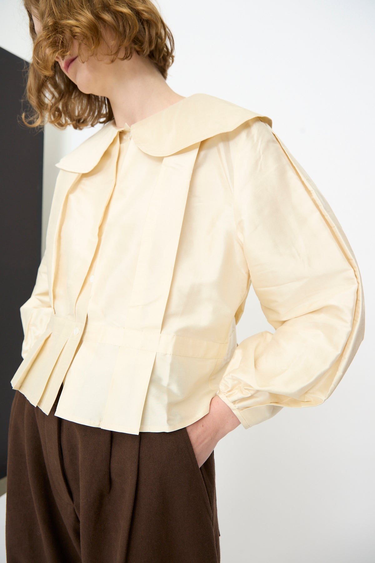 A person stands against a plain background with one hand in their pocket, wearing brown pants and the Cawley Silk Dupion Box Pleat Blouse in Ivory. This elegant long-sleeve blouse, featuring a large collar and crafted in the UK, adds a timeless touch to the outfit.
