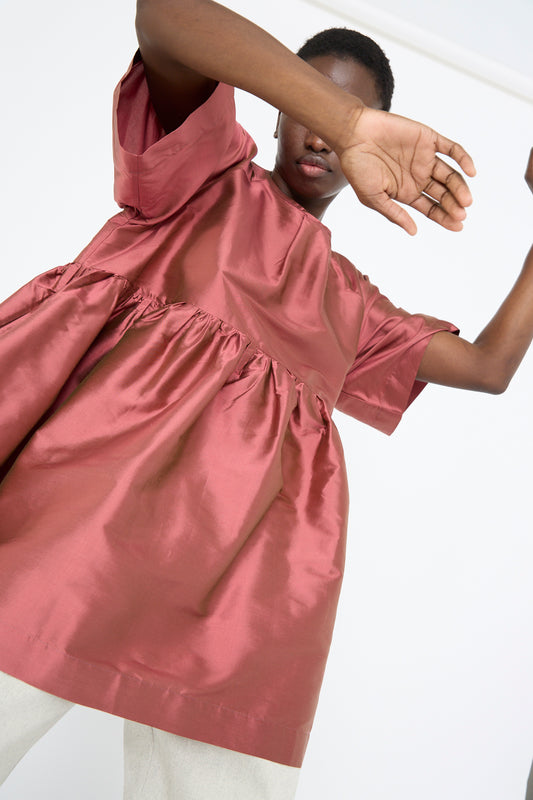 A person in Cawley's Silk Dupion Delilah Dress in Rhubarb, featuring a crew neckline and an oversized mini dress style, poses with one hand raised to obscure part of their face. White pants are partially visible below.