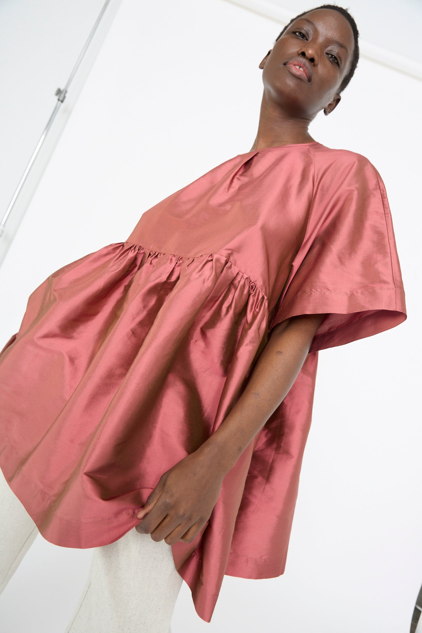 A person in a silk Dupion Delilah Dress by Cawley in rhubarb, featuring a loose fit and crew neckline, gazes slightly upwards against a neutral background.