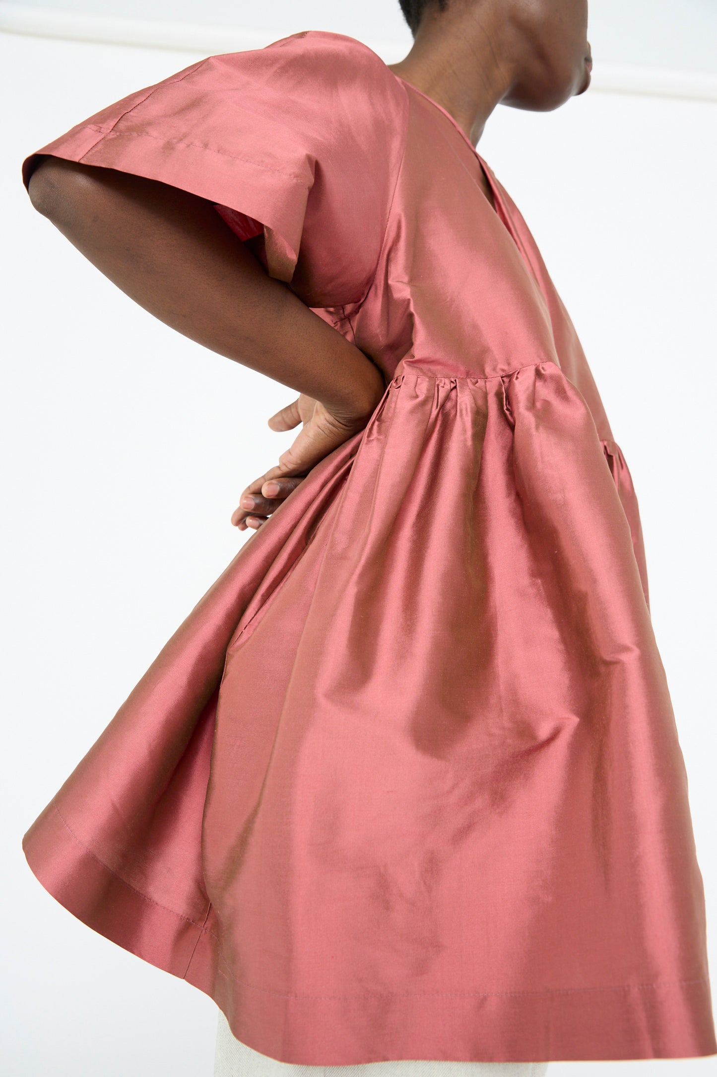 A person wearing Cawley's Silk Dupion Delilah Dress in Rhubarb with a crew neckline and short sleeves poses hands on hips against a plain background.