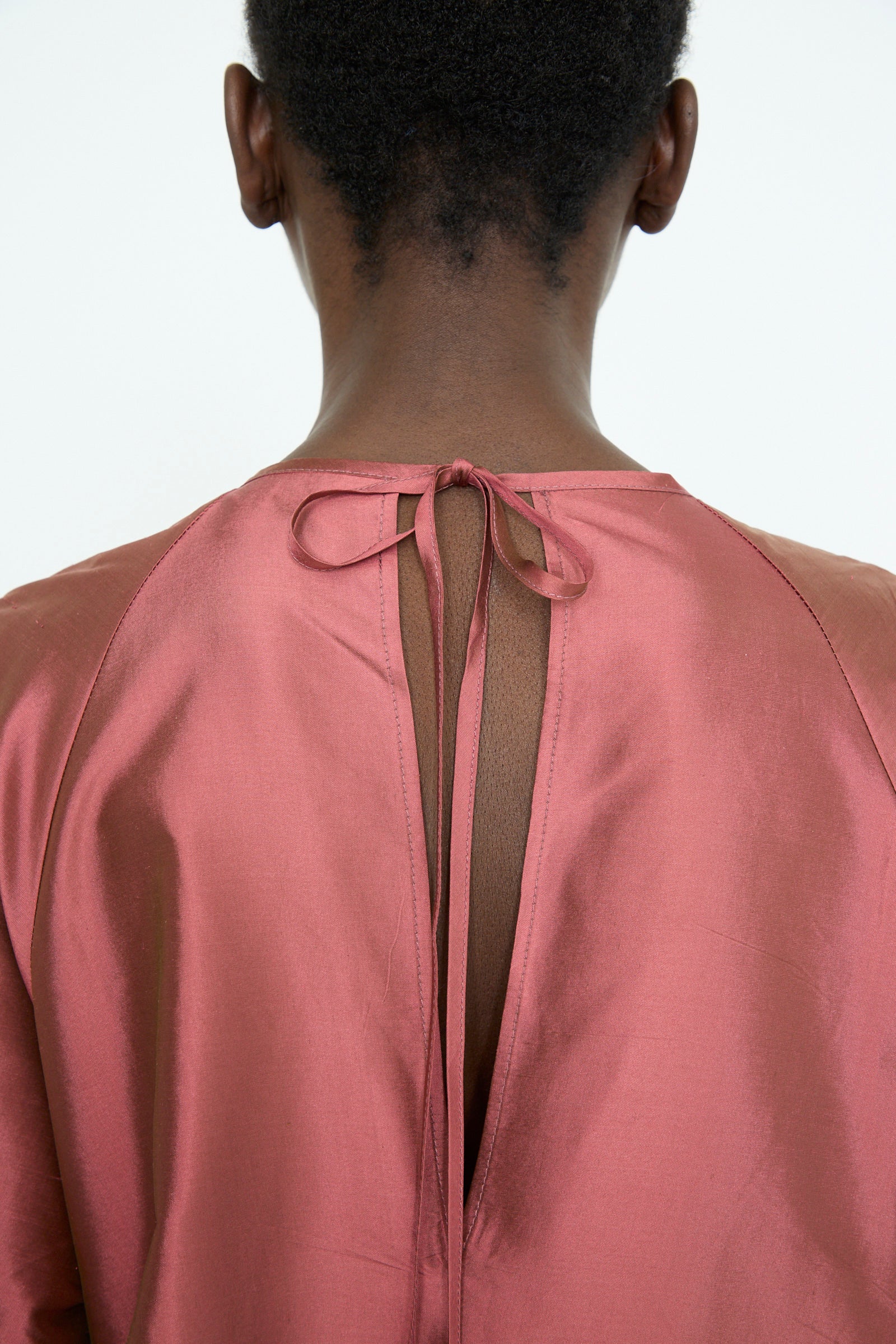 A person is seen from the back wearing an oversized Silk Dupion Delilah Dress in Rhubarb by Cawley. The dress is made of lightweight silk, featuring an open back and a tie closure at the neckline.