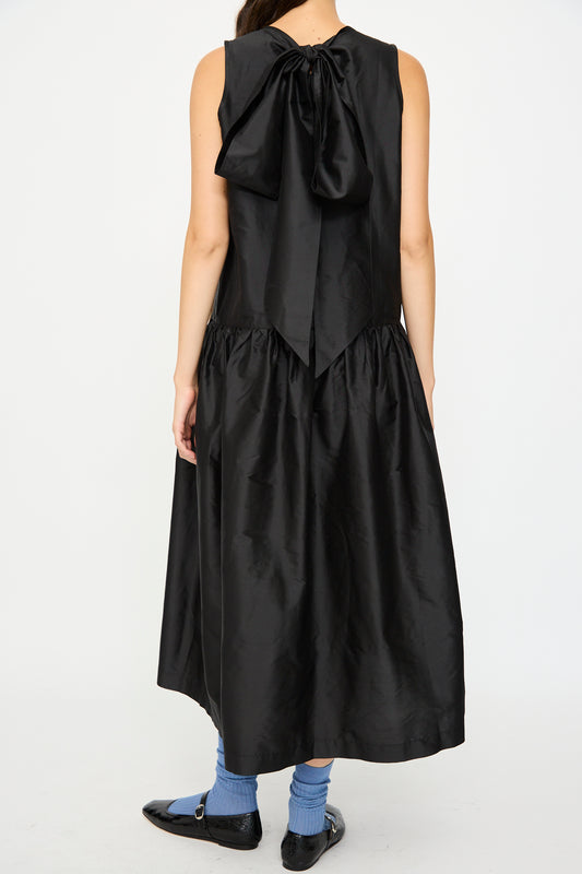 A person wearing the Cawley Silk Dupion Gathered Isabella Dress in Black, blue socks, and black shoes stands against a white background.