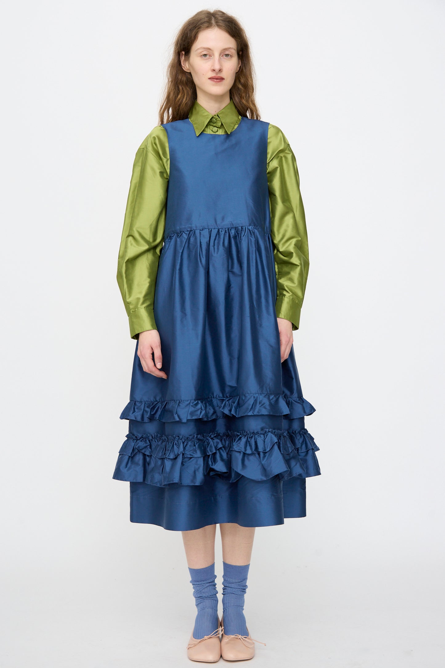A person wears Cawley’s Silk Dupion Genevieve Dress in Insignia, styled over a green satin shirt with blue socks and beige flats, standing against a plain background.