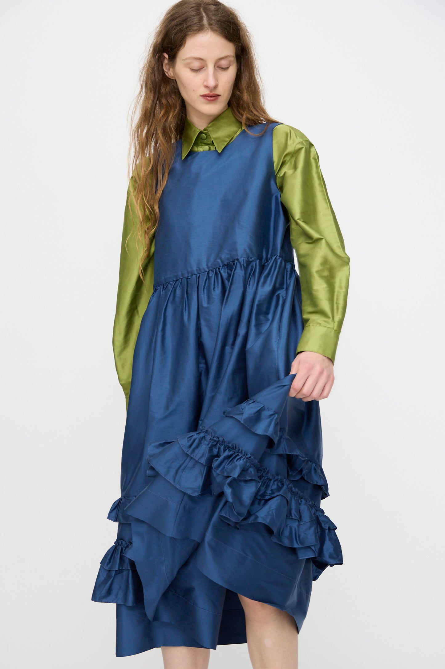 Wearing Cawley’s Silk Dupion Genevieve Dress in Insignia, made in London, a person pairs it with a shiny green long-sleeve shirt underneath and looks down while holding the ruffled dress.
