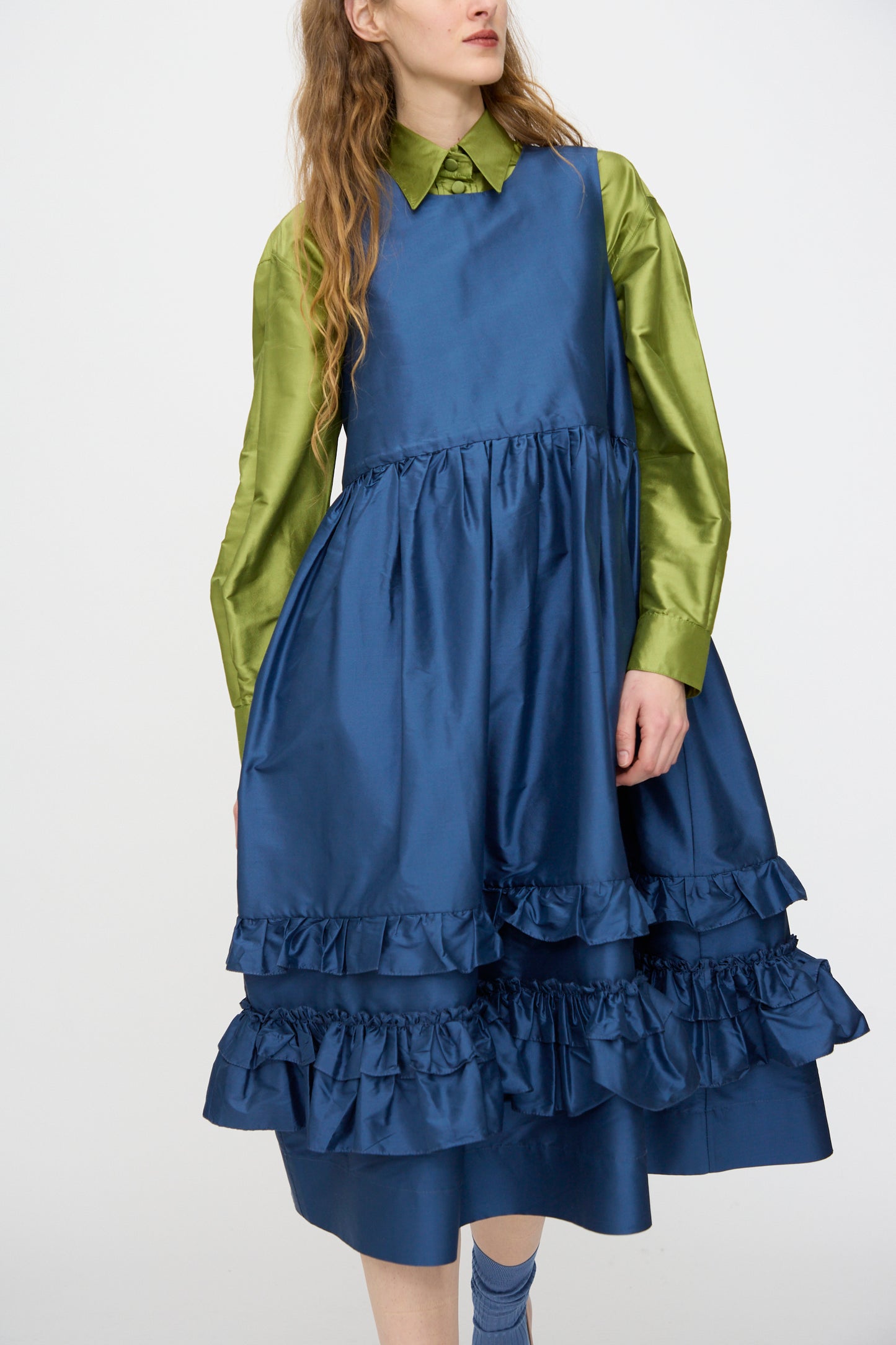 A person wears the Cawley Silk Dupion Genevieve Dress in Insignia, a sleeveless blue ruffled design made in London, layered over a shiny green long-sleeve shirt and set against a plain light background.