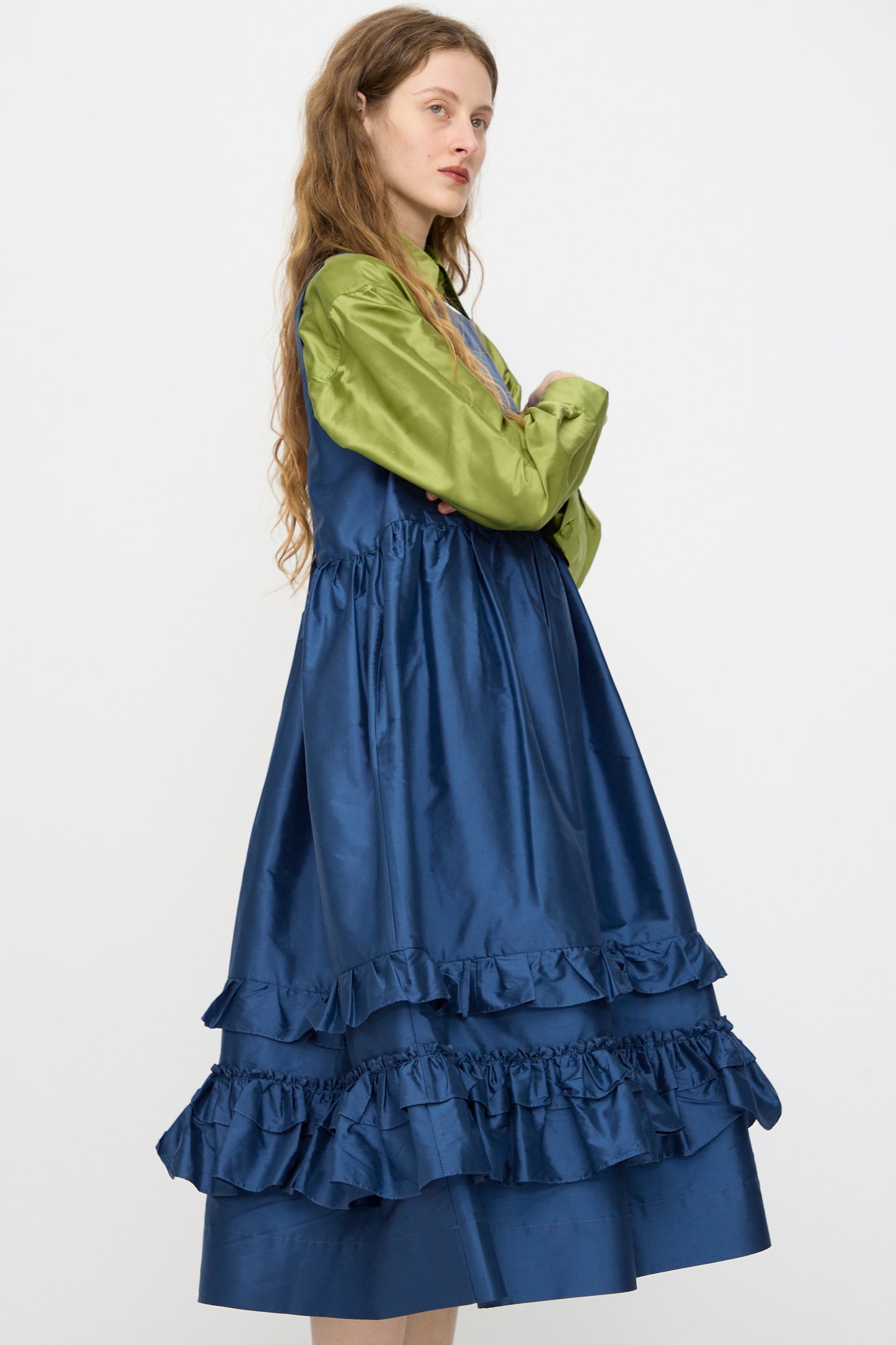A woman with long wavy hair and a neutral expression wears the Cawley Silk Dupion Genevieve Dress in Insignia, layered over a light green long-sleeve shirt, standing sideways against a plain white background.