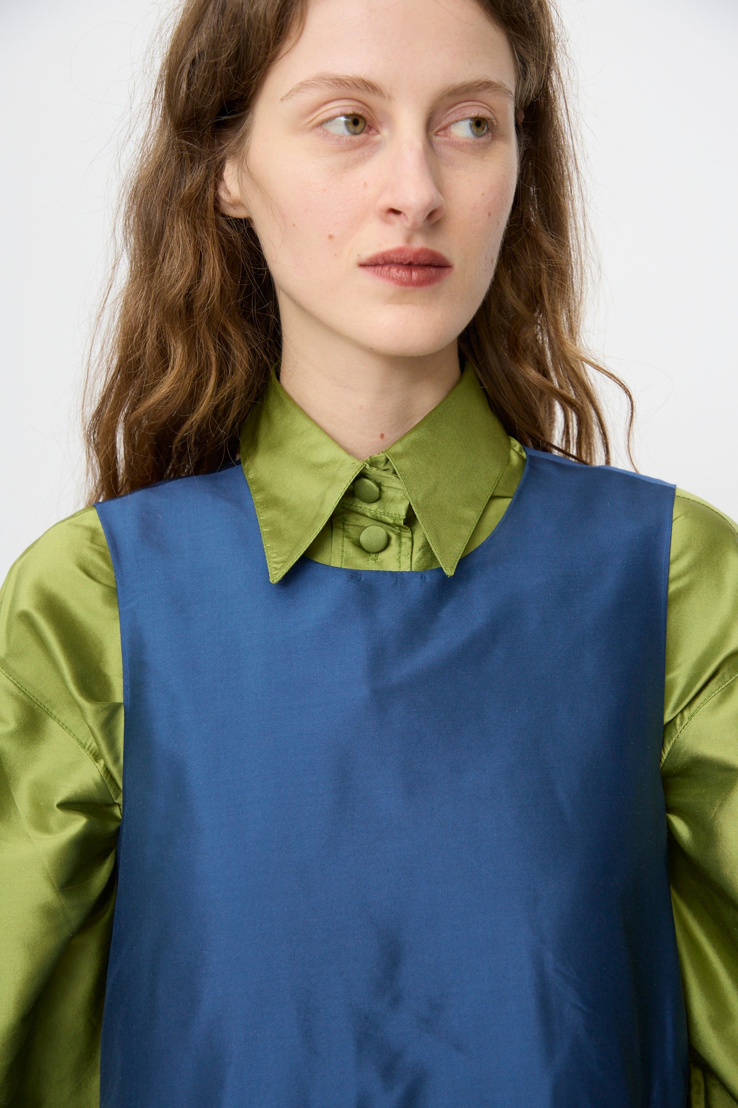 A woman with long brown hair wears the Cawley Silk Dupion Genevieve Dress in Insignia over a shiny green collared shirt, gazing to the side against a plain backdrop. 