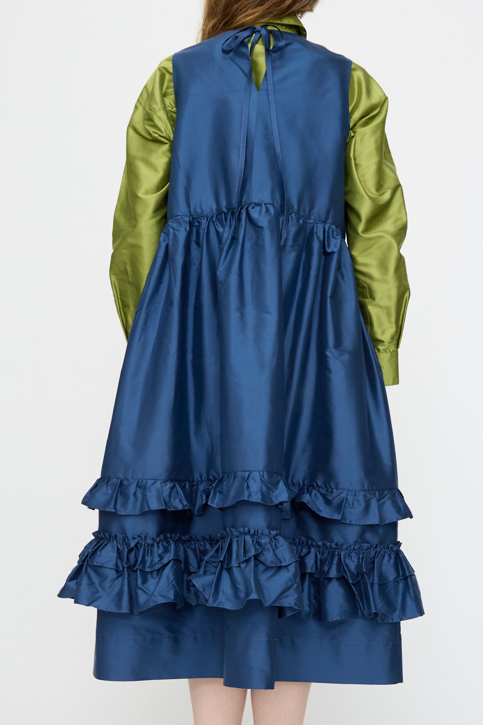 A person is seen from behind wearing the Cawley Silk Dupion Genevieve Dress in Insignia—a shiny blue, ruffled sleeveless dress made in London—over a long-sleeve green shirt, set against a plain background.