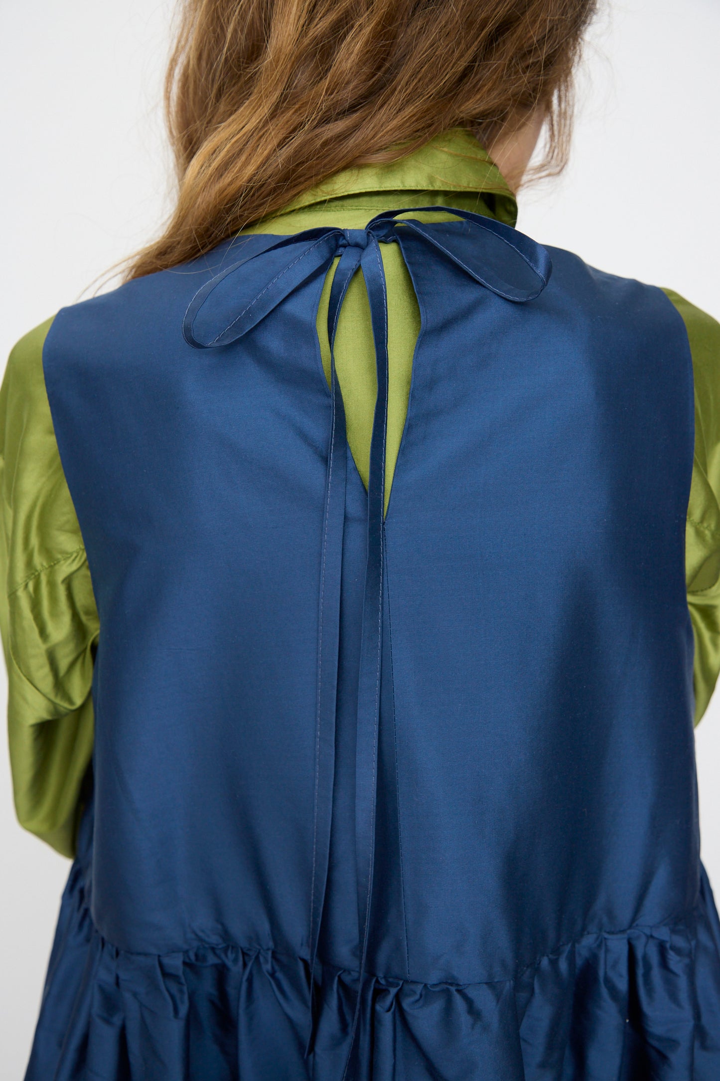 A person with long brown hair wears the Cawley Silk Dupion Genevieve Dress in Insignia, a sleeveless blue midi dress made in London, styled over a shiny green shirt and tied at the back.