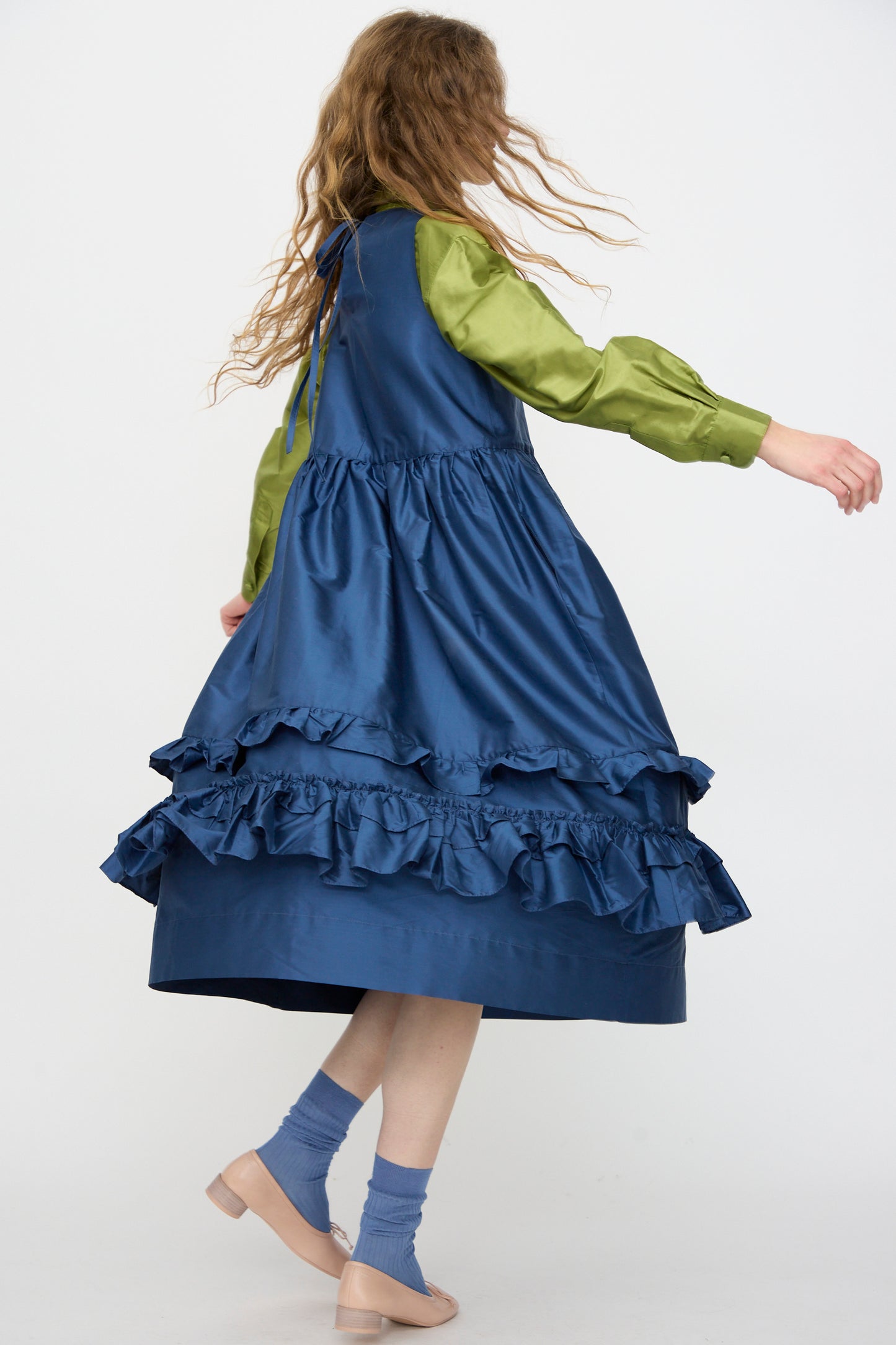 A person with long wavy hair, seen from behind while walking, wears a green long-sleeve top under the Cawley Silk Dupion Genevieve Dress in Insignia, with blue socks and beige shoes.