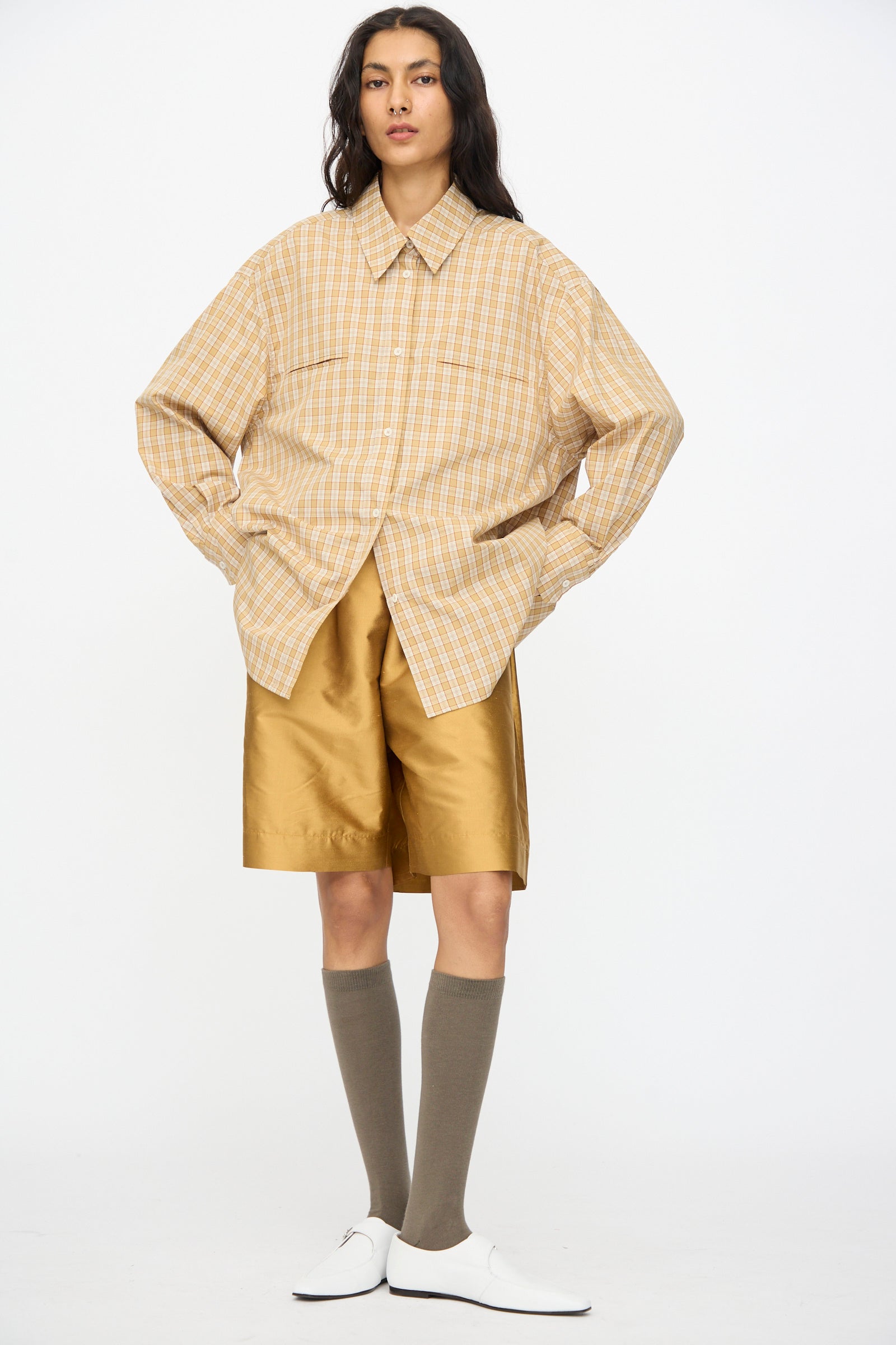 A person stands in a loose plaid shirt, Cawley Silk Dupion Hilda Short in Ochre with an elastic waistband, knee-high brown socks, and white shoes against a plain white background.