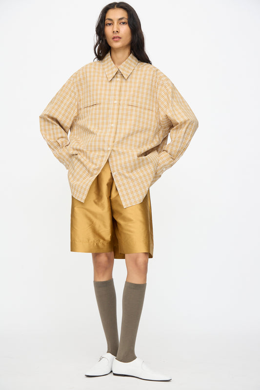A person stands in a loose plaid shirt, Cawley Silk Dupion Hilda Short in Ochre with an elastic waistband, knee-high brown socks, and white shoes against a plain white background.