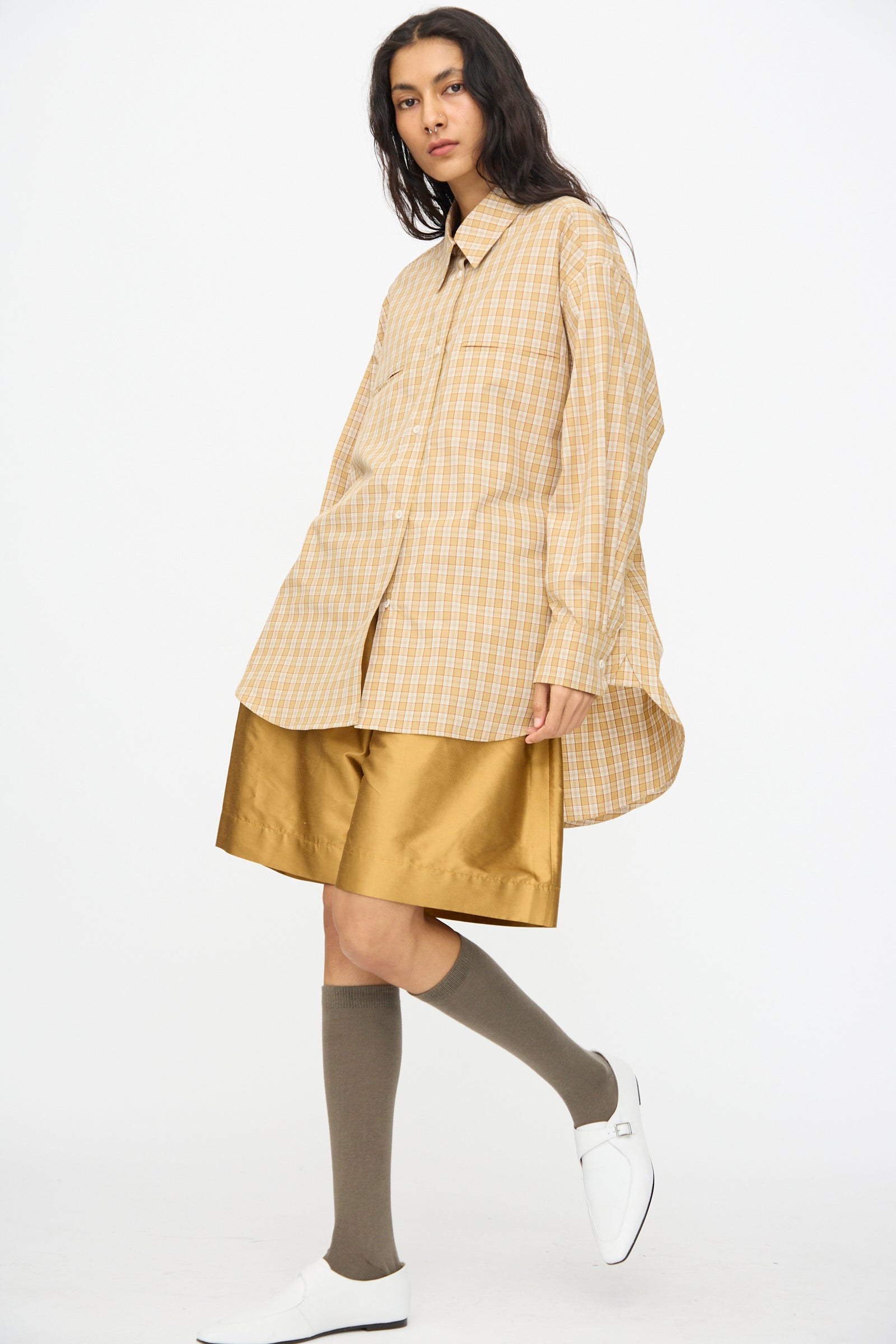A person wearing a loose beige plaid shirt, Cawley Silk Dupion Hilda Short in Ochre, taupe knee-high socks, and white shoes stands against a plain white background.