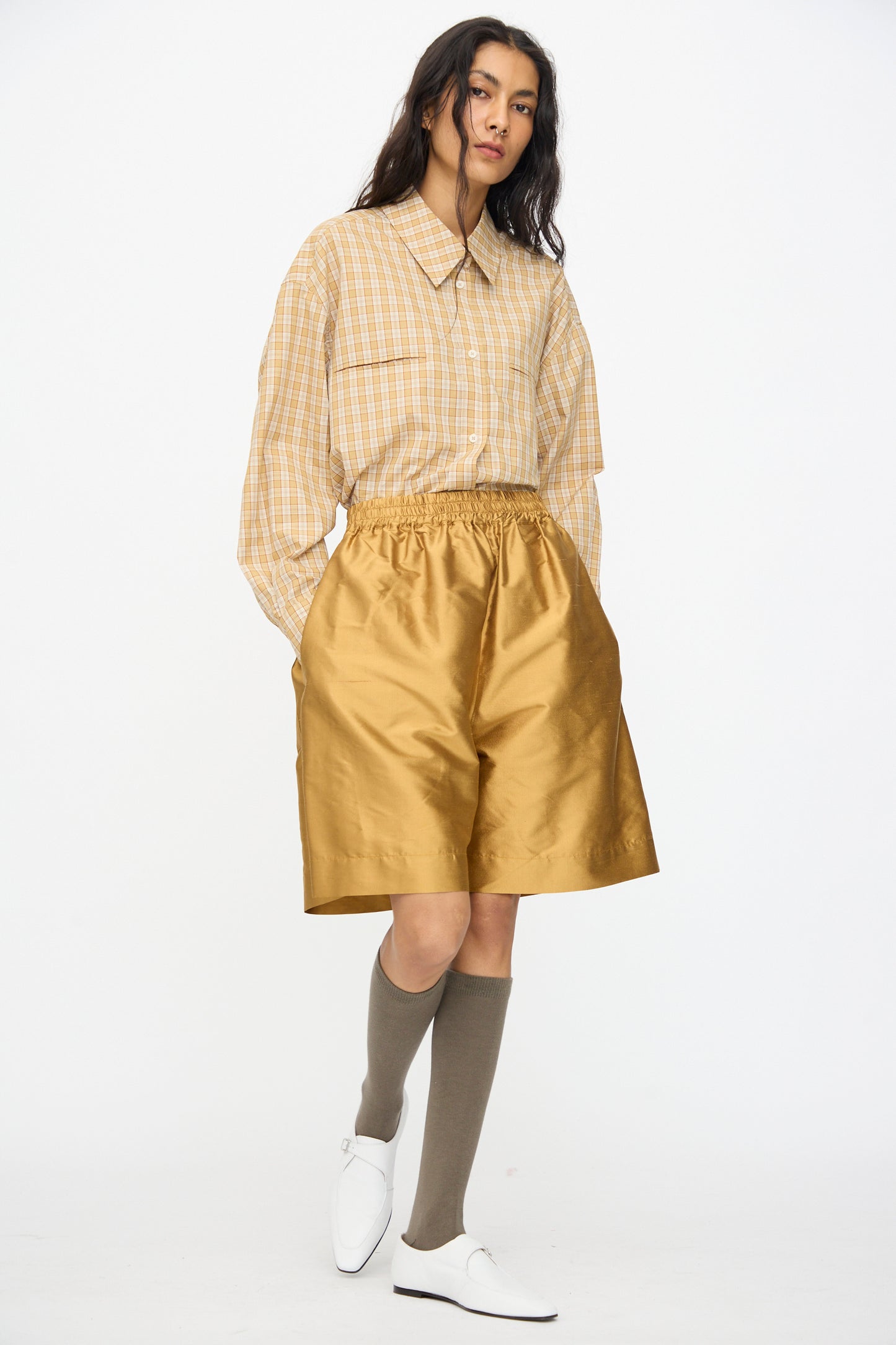 A person wears the Cawley Silk Dupion Hilda Short in Ochre with a beige plaid shirt, knee-high gray socks, and white shoes, standing against a plain white background.