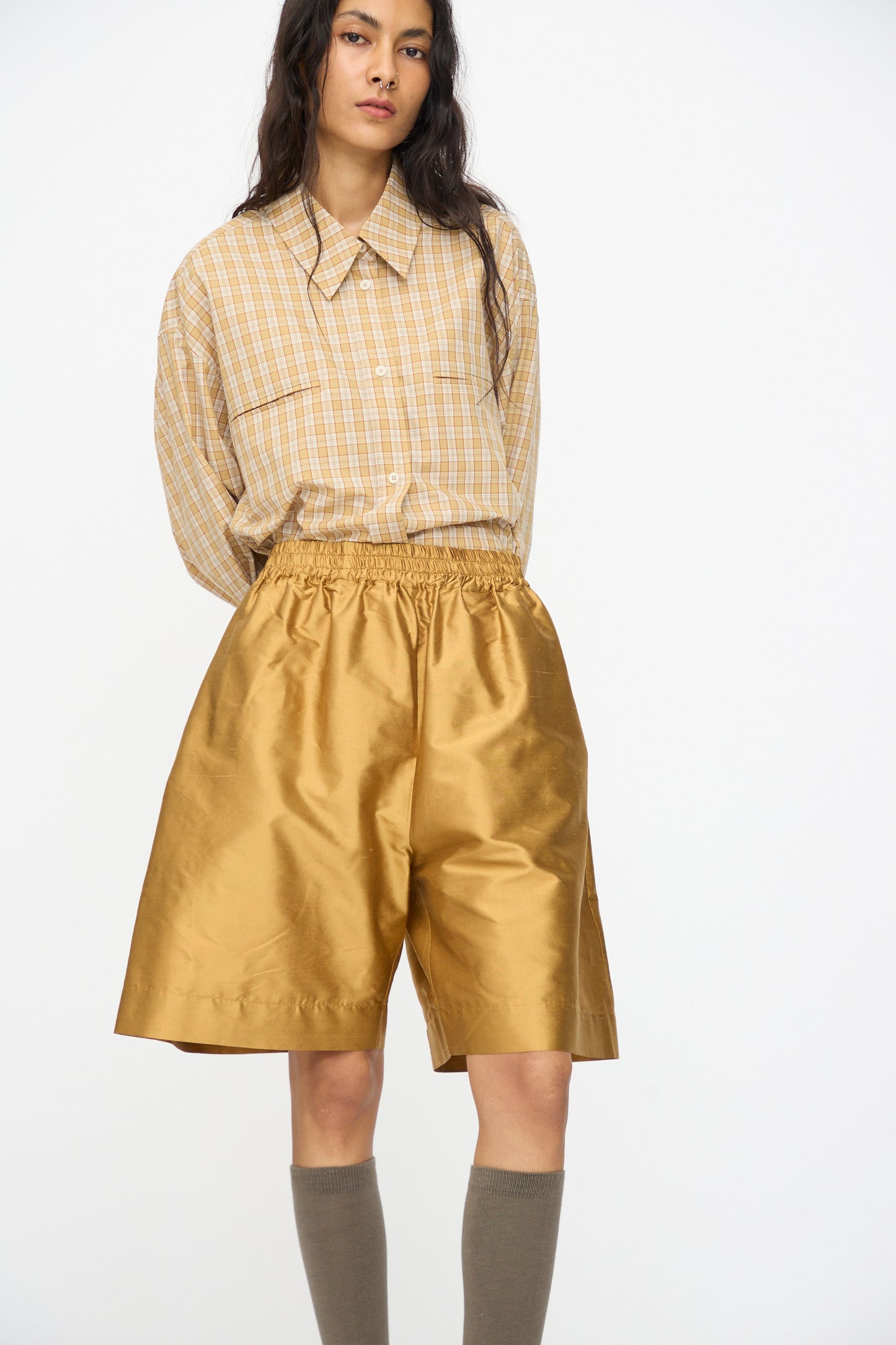A person wearing a beige plaid shirt, Cawley’s Silk Dupion Hilda Short in Ochre, and knee-high taupe socks stands against a plain white background.
