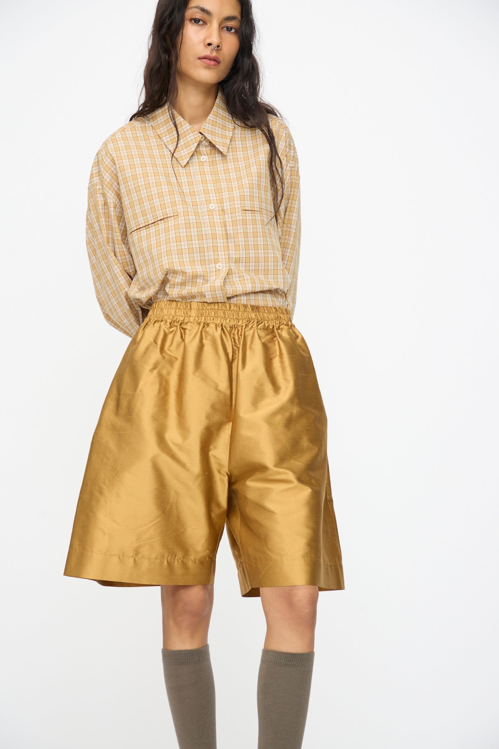 A person wearing a beige plaid shirt, Cawley’s Silk Dupion Hilda Short in Ochre, and knee-high taupe socks stands against a plain white background.