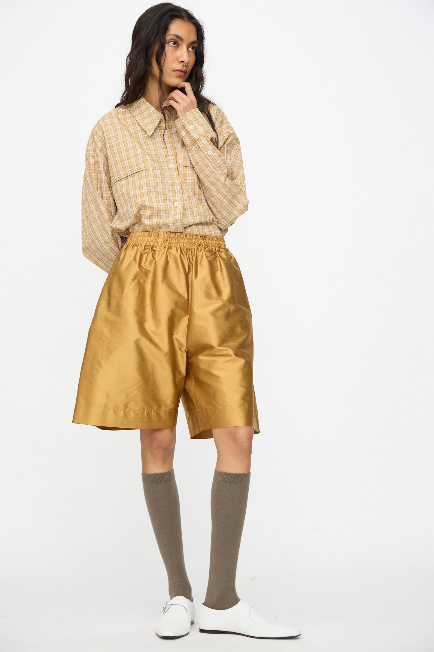A person wears a beige plaid shirt tucked into Cawley's Silk Dupion Hilda Short in Ochre, paired with knee-high taupe socks and white shoes, against a plain white background. 