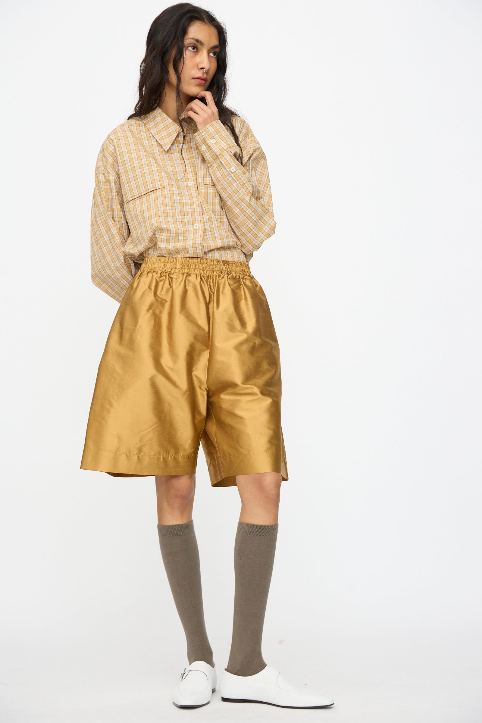 A person wears a beige plaid shirt tucked into Cawley's Silk Dupion Hilda Short in Ochre, paired with knee-high taupe socks and white shoes, against a plain white background. 