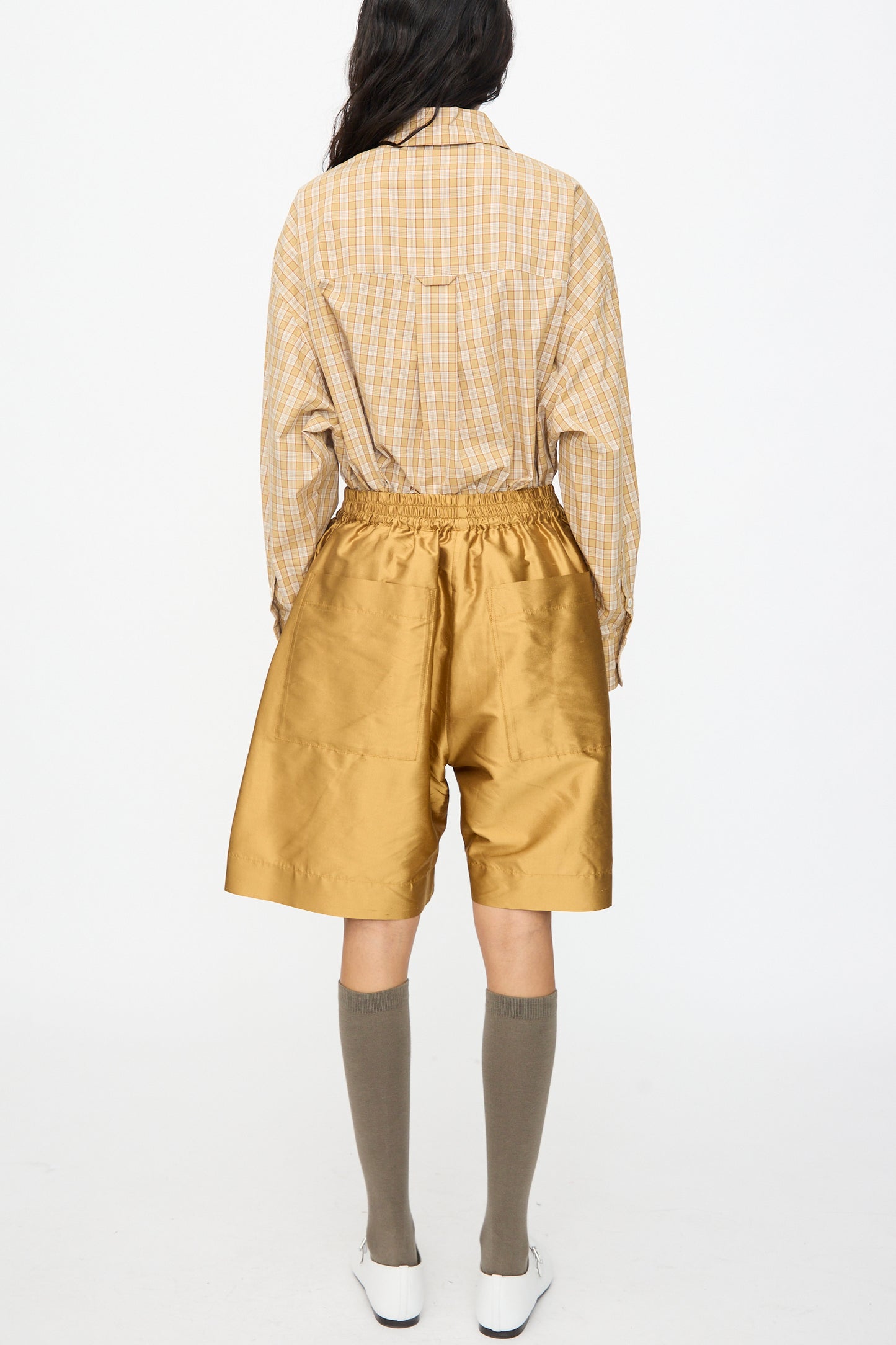 A person stands facing away in a beige plaid shirt tucked into Cawley's Silk Dupion Hilda Short in Ochre, featuring an elastic waistband, knee-high gray socks, and white shoes, against a plain white background. Made in London.
