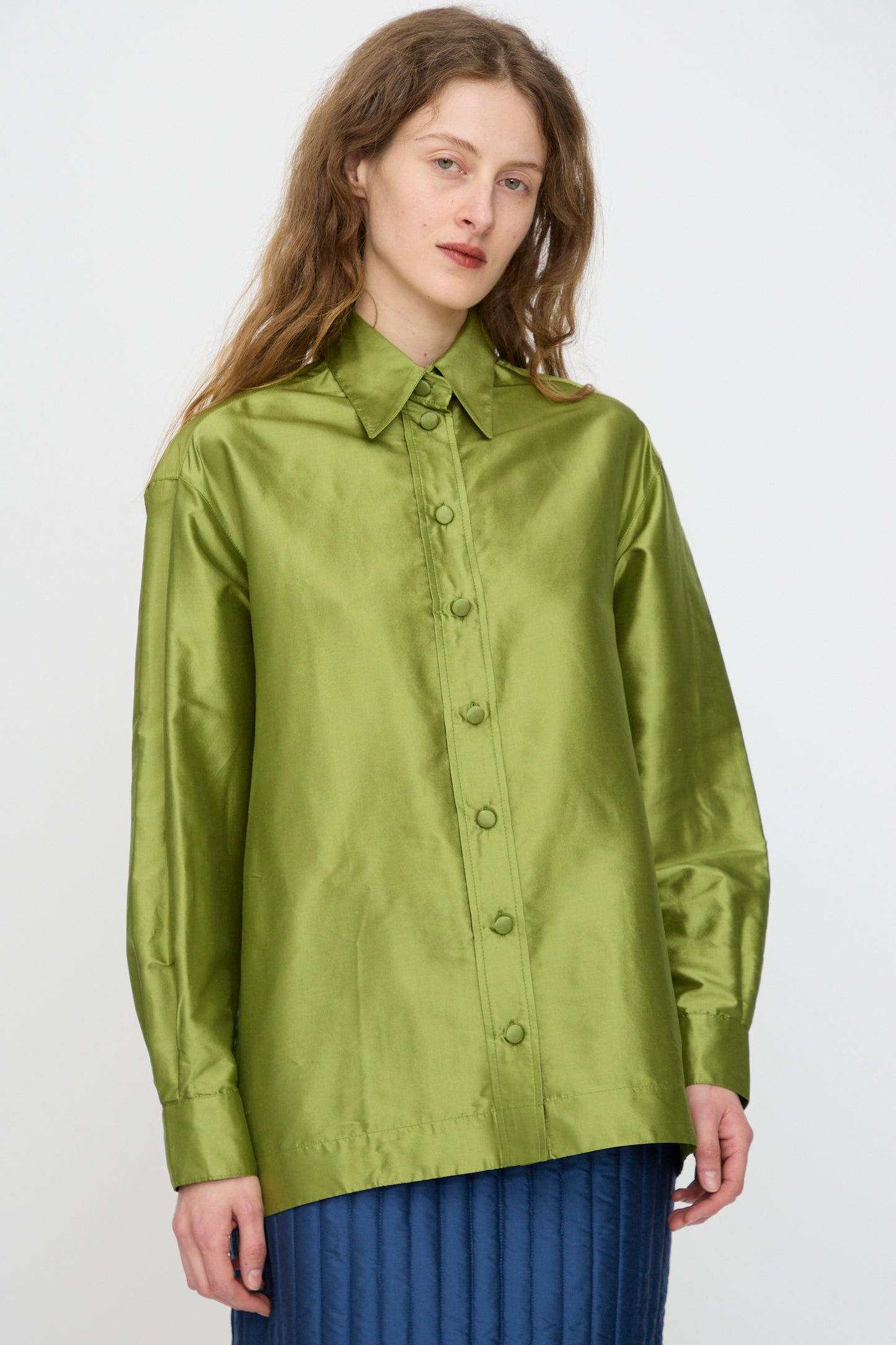 A woman wears the Cawley Silk Dupion June Shirt in Apple, a loose-fitting, shiny green long-sleeve top, paired with a blue quilted skirt, standing against a plain light background.