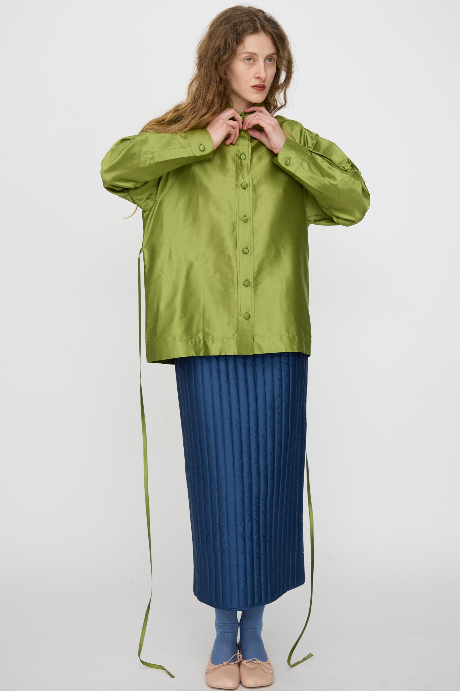 A person ties the strings of a Cawley Silk Dupion June Shirt in Apple—a shiny green silk top—paired with a long, quilted blue skirt, set against a plain white background.