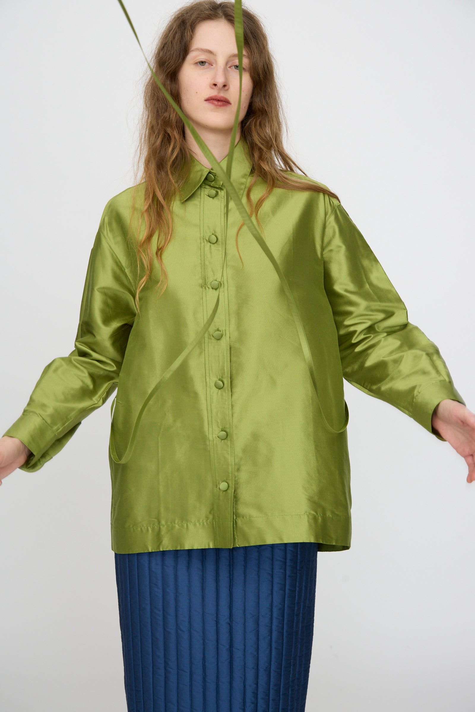A woman wears the Cawley Silk Dupion June Shirt in Apple and a blue quilted skirt, standing against a plain white background with a green plant stem partially covering her face.