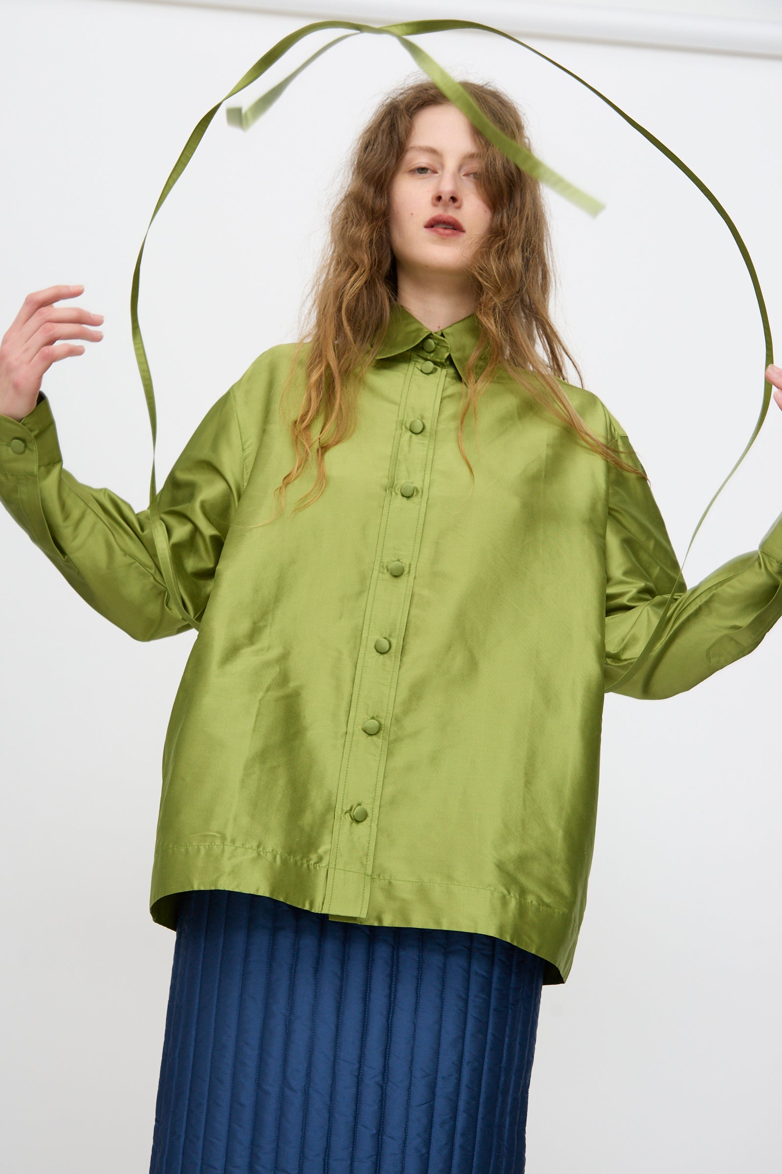 A woman wears the Cawley Silk Dupion June Shirt in Apple, arms raised as she holds a green ribbon above her head, against a plain background.