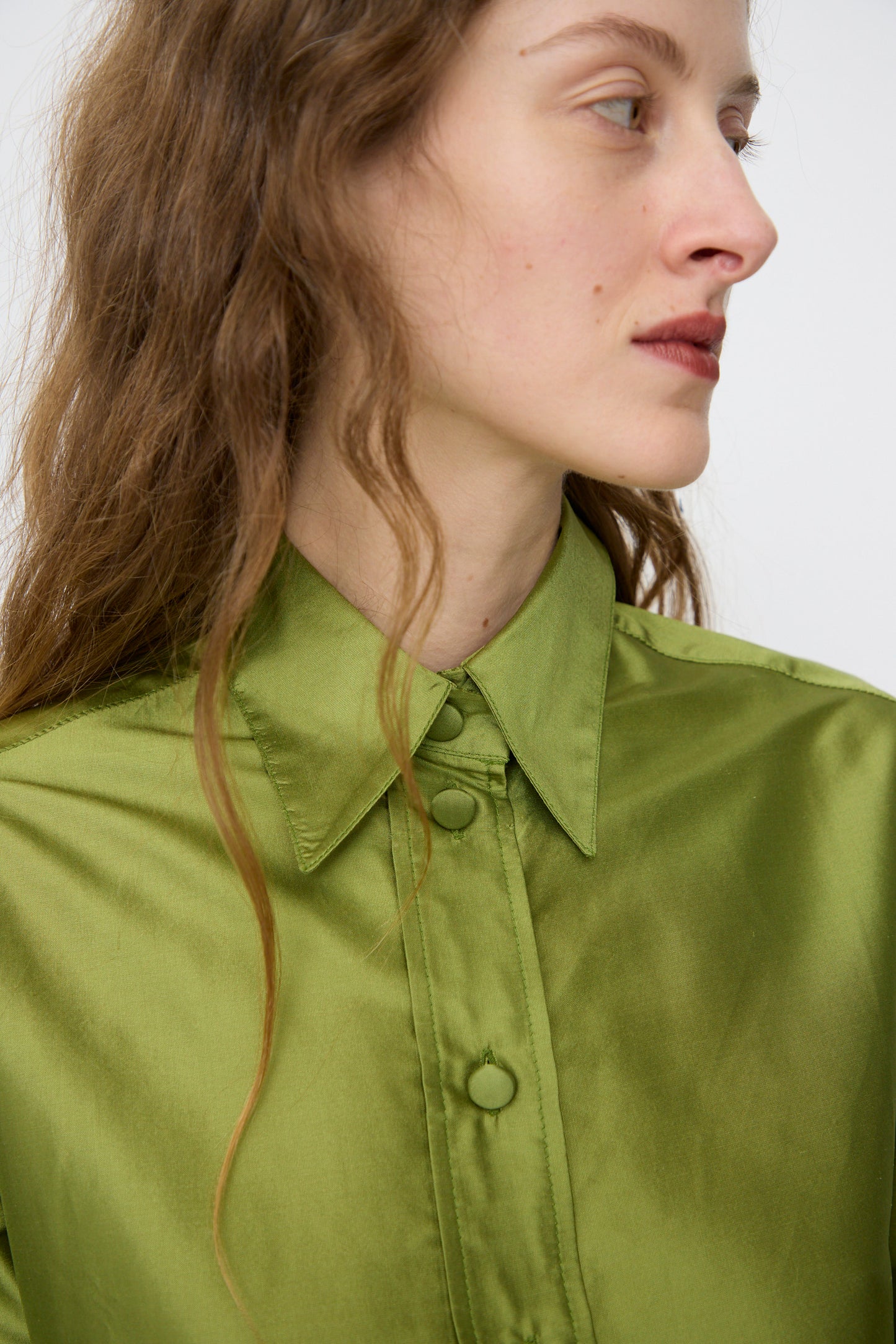 A person with long brown hair wears the Cawley Silk Dupion June Shirt in Apple, looking to the side against a plain background.