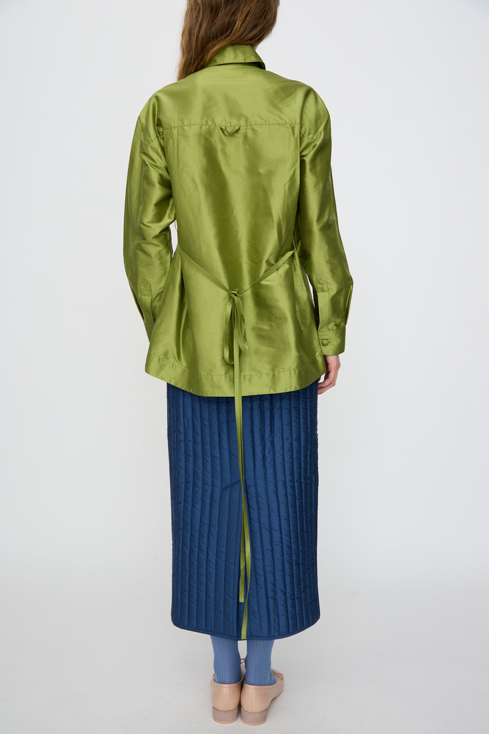 A person shown from the back wears the Cawley Silk Dupion June Shirt in Apple, paired with a quilted blue skirt, socks, and beige shoes against a plain white background.