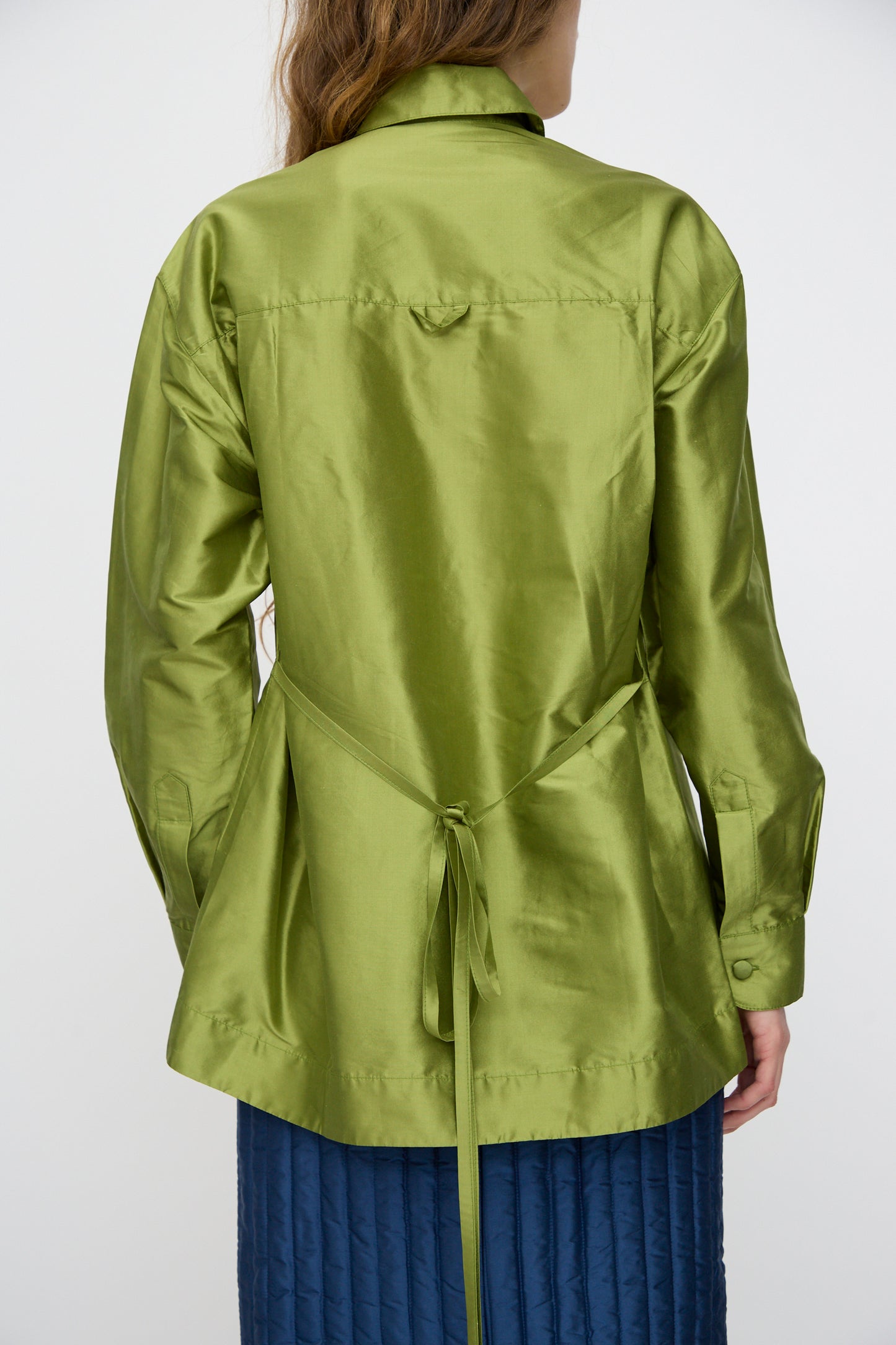 Someone wears the Cawley Silk Dupion June Shirt in Apple—a shiny green silk button-up with a tie detail at the back—paired with a blue quilted skirt. The photo shows the outfit from behind.