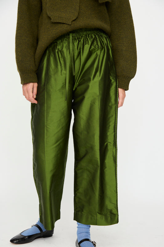 A person stands against a plain white background, wearing Cawley's Silk Dupion Luna Trouser with Trim in Bottle, paired with a dark green sweater, blue socks, and black shoes.